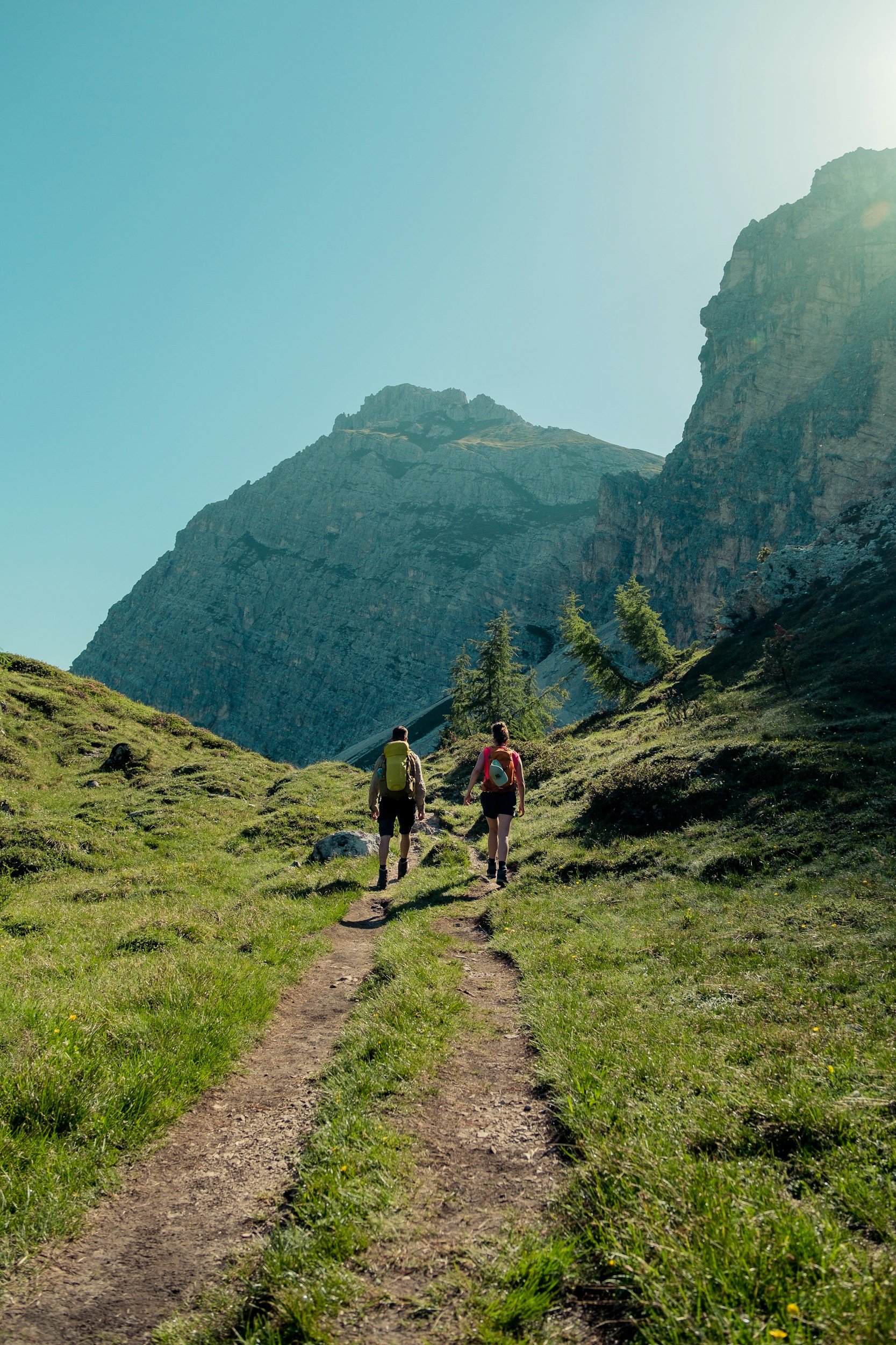 Two hikers walking along a dirt trail through a grassy mountain landscape with large rocky cliffs towering in the background under a clear blue sky.
