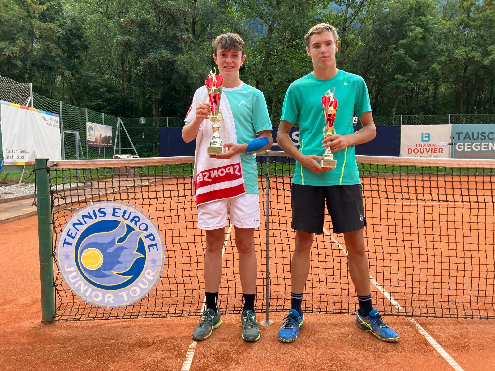 Zwei Jugendliche stehen auf einem Tenniscourt hinter dem Netz, jeder hält einen Pokal. Im Hintergrund Bäume und Werbetafeln.