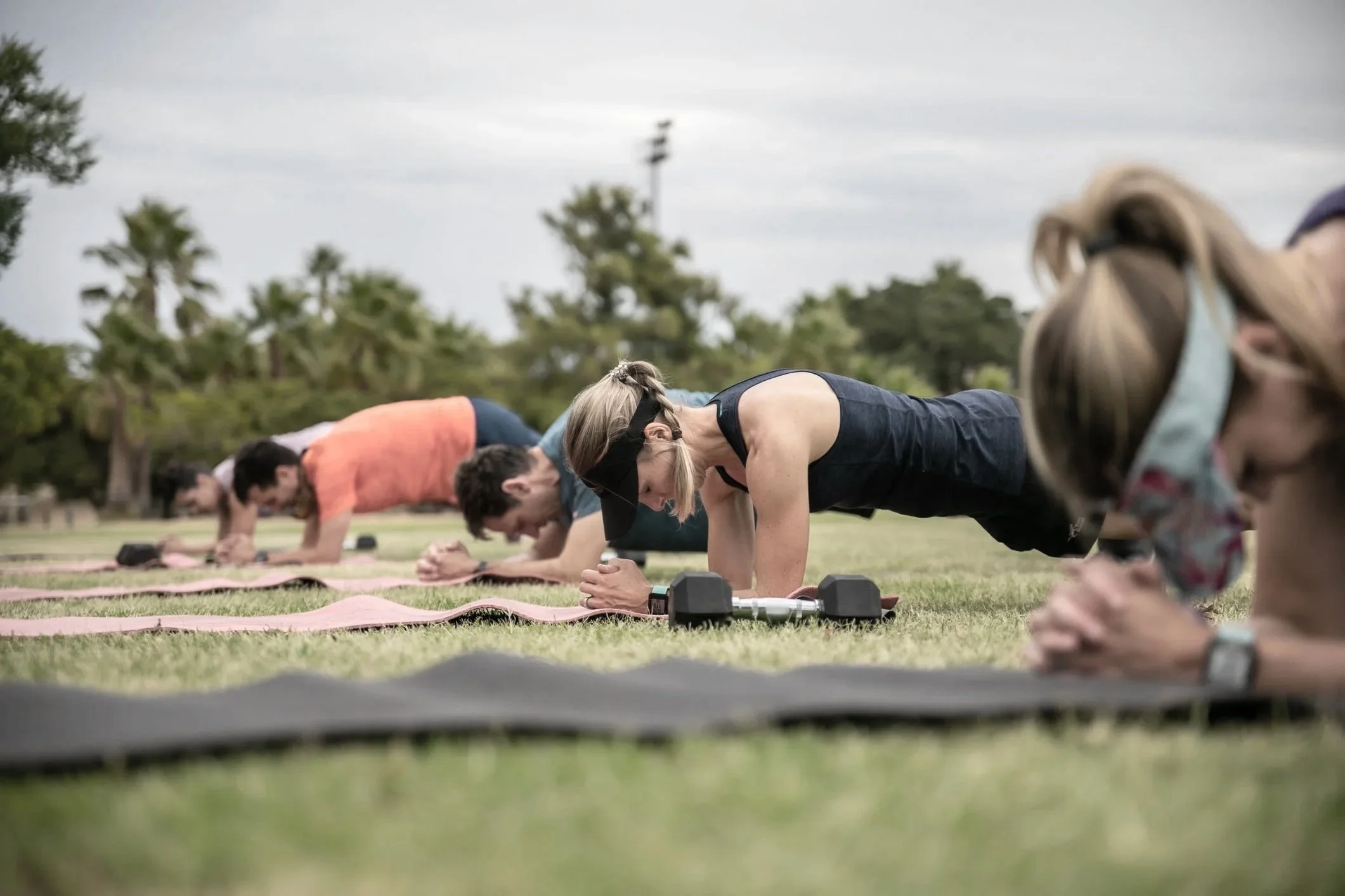 Menschen beim Yoga im Freien, in einer Reihe im Cobra- und Unterarmstütz-Position, mit Hanteln und Yoga-Matten auf einer Wiese.