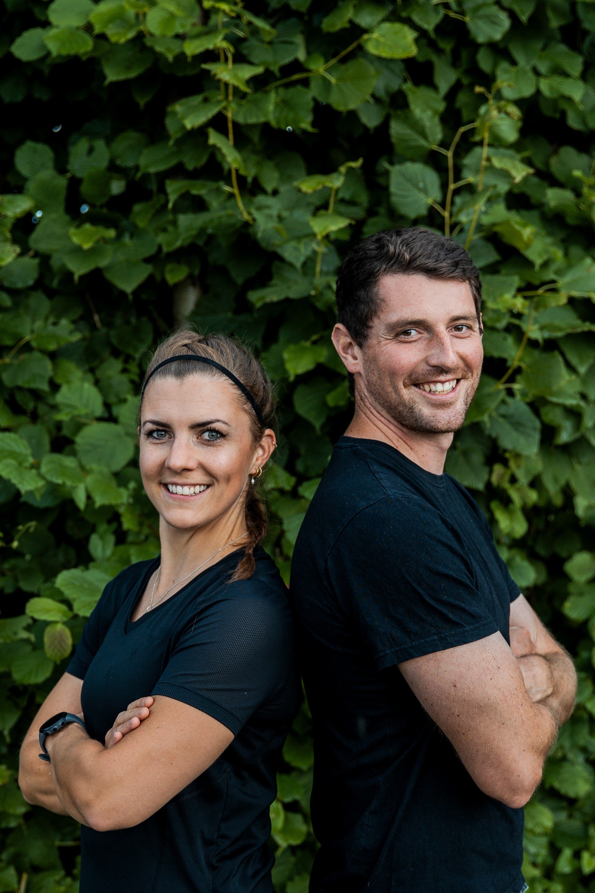 Ein Mann und eine Frau stehen Rücken an Rücken vor einer grünen Blätterwand, lächeln und schauen in die Kamera, beide tragen schwarze T-Shirts.