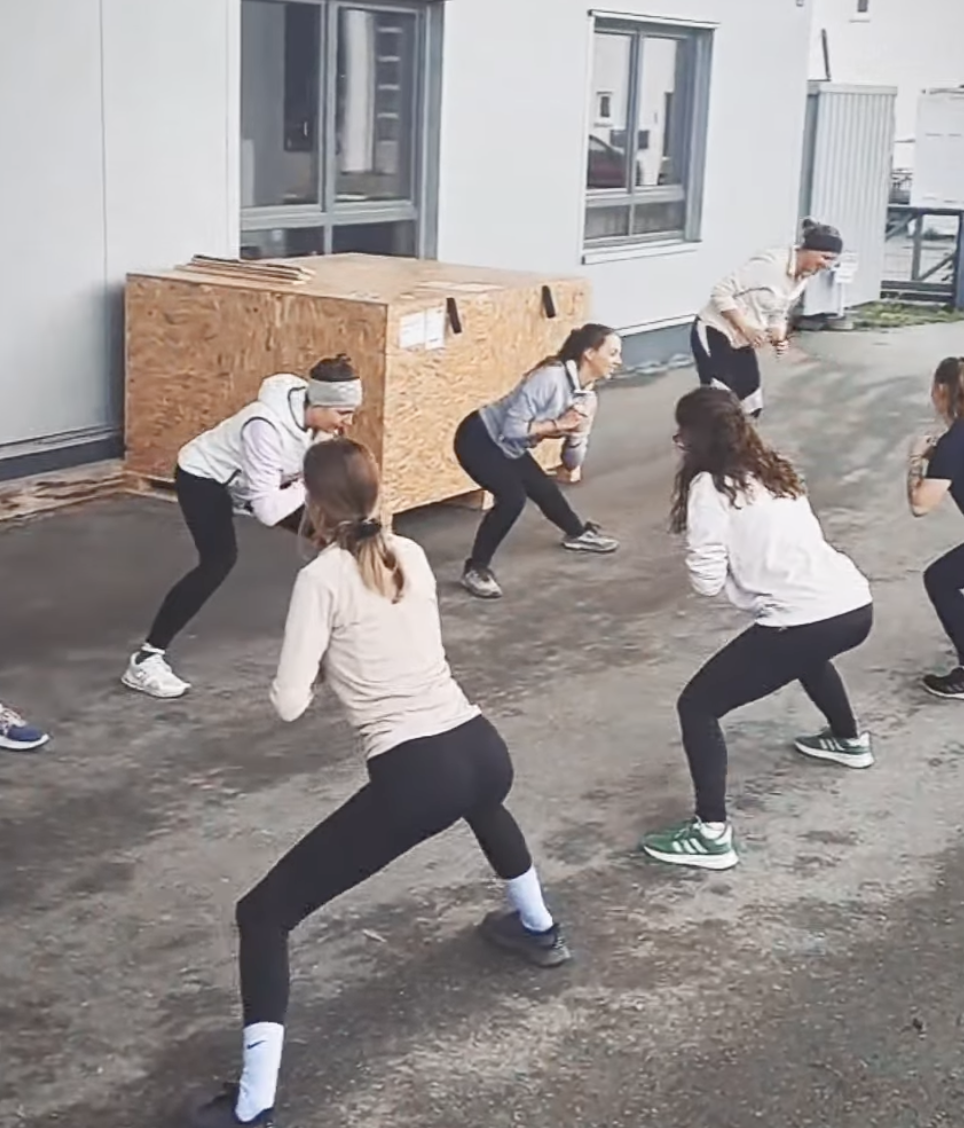 Menschen, die in einer Gruppe draußen Übungen machen, die Knie beugen und springen, während ein Raum im Hintergrund sichtbar ist.
