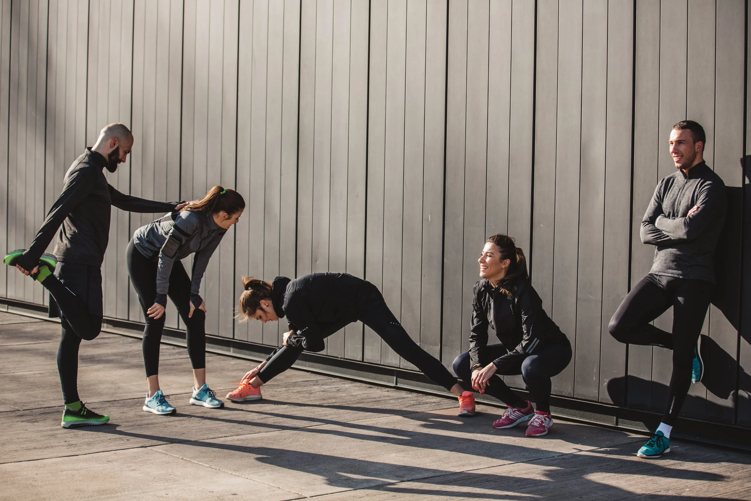 Fünf Menschen, die sich draußen vor einer modernen Holzwand dehnen und auf ein Workout vorbereiten, mit lachender Frau in der Mitte, während zwei Männer stehen, einer mit Hanteln, die anderen sitzen gegen die Wand.