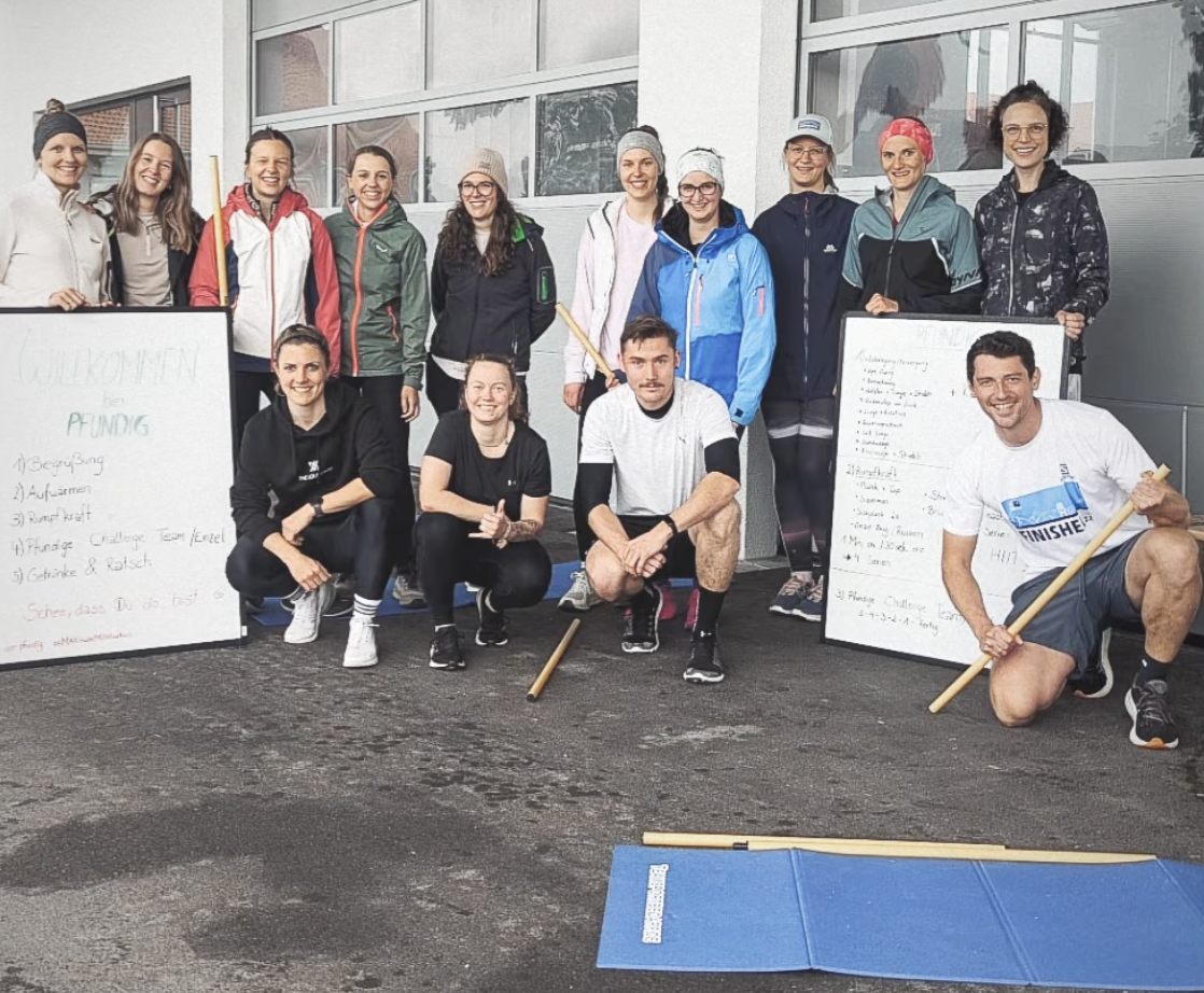 Gruppe von Menschen in Sportkleidung, die vor einem Gebäude posieren, einige halten Schilder mit Text, im Vordergrund stehen drei Menschen mit Holzstäben, alle lachen und scheinen nach einem Lauf-Event oder einem sportlichen Wettbewerb zusammen zu sein.