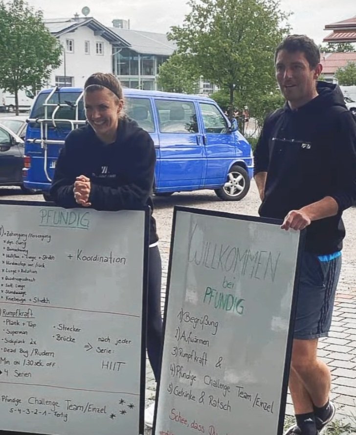 Zwei Personen mit Whiteboards im Freien, die für ein Sportevent oder Training vorbereitet sind, mit einem blauen Van im Hintergrund.