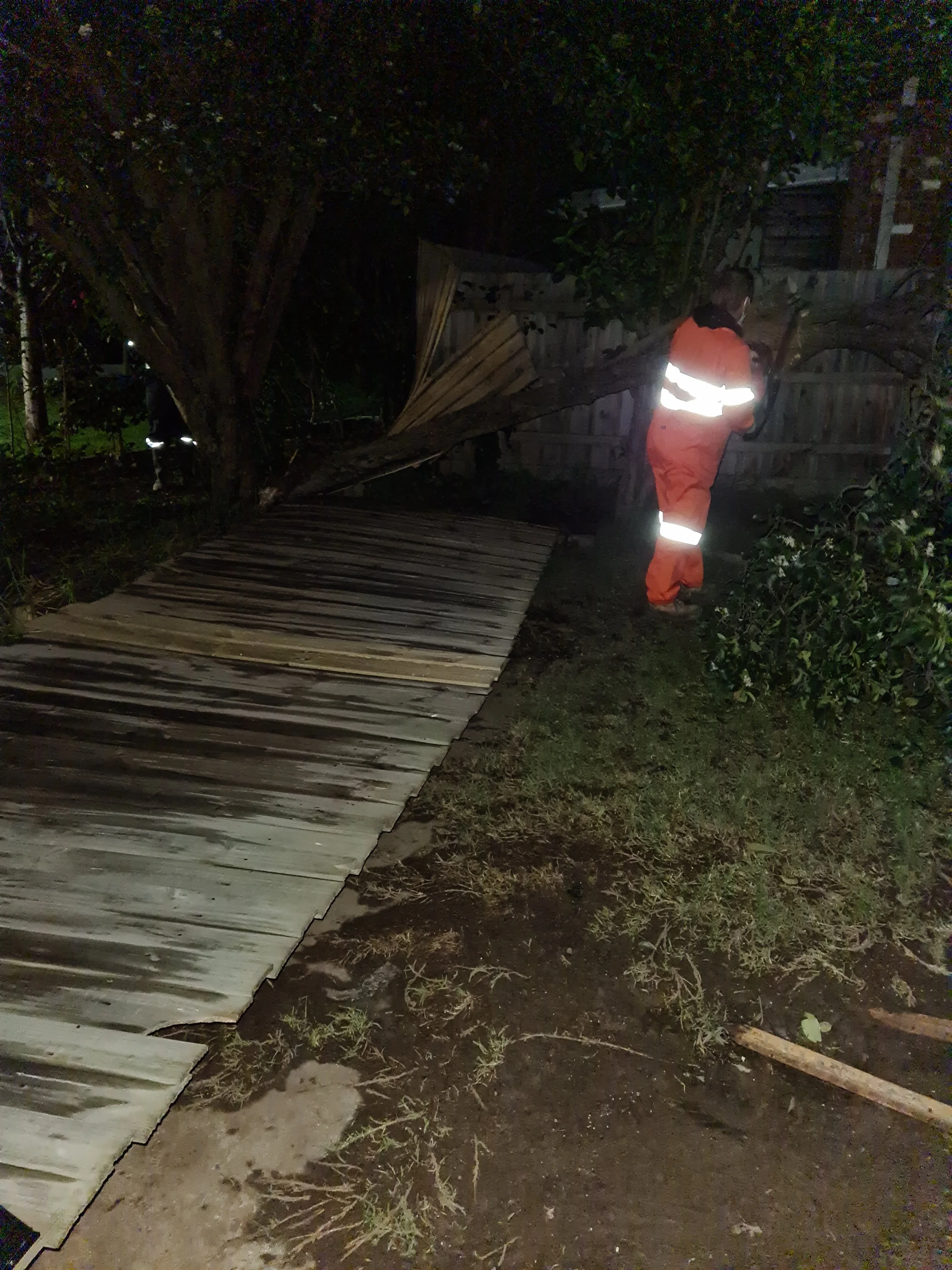 Nighttime scene of a person in an orange safety jumpsuit inspecting a fallen tree on a damaged wooden fence with a broken gate.