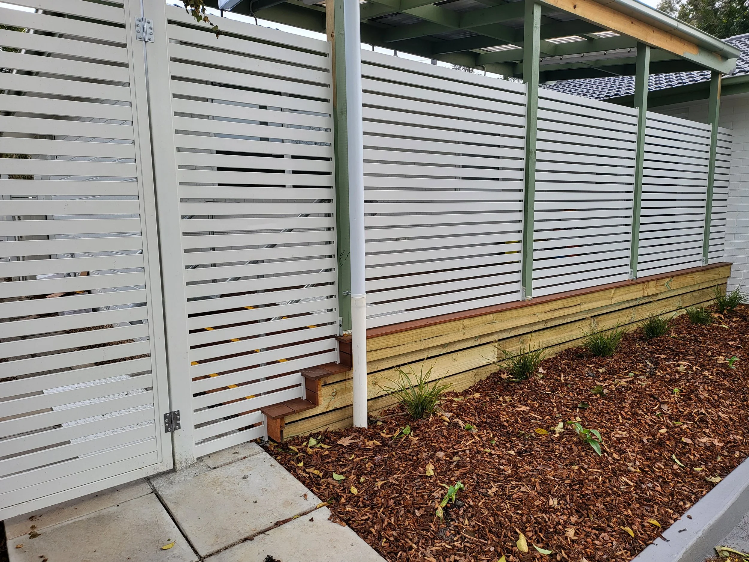 A white horizontal slatted fence with a small gate, attached to a white brick house with a dark-colored tiled roof, surrounded by a garden with brown mulch and small green plants.
