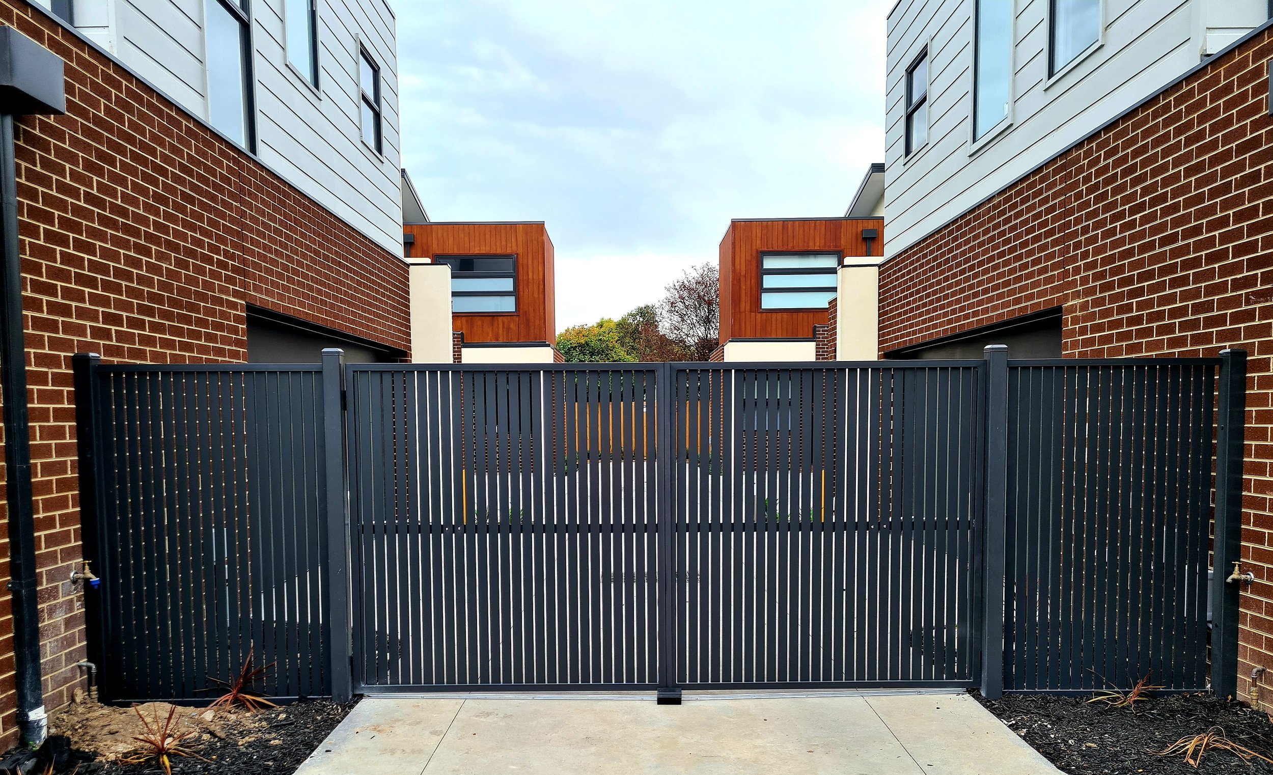 View of a black metal gate between two modern apartment buildings with brick and wood siding, and windows in the background.