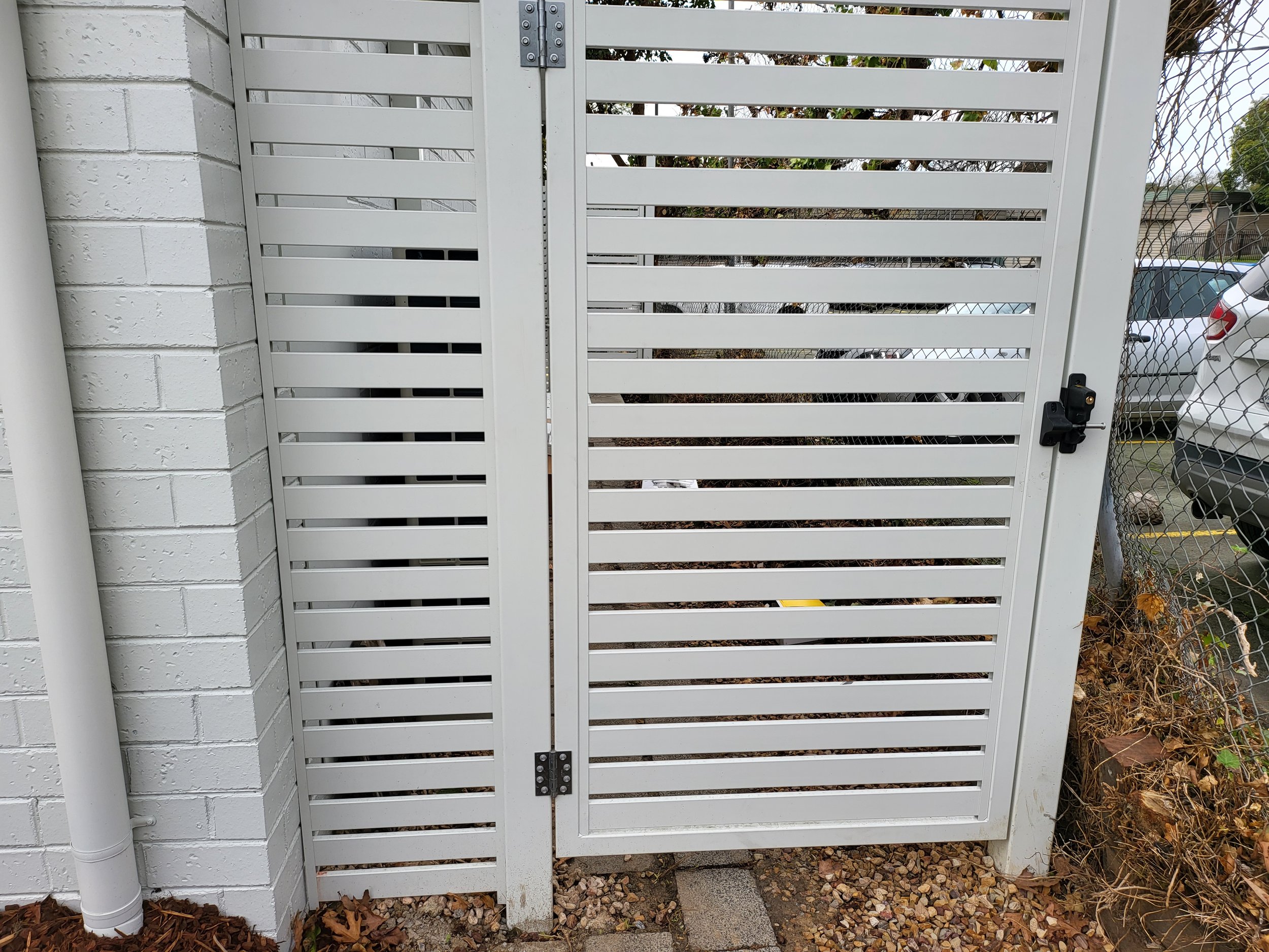 A white metal facility gate with horizontal slats, secured with a black latch, located next to a white brick wall, with parked cars visible on the right side.