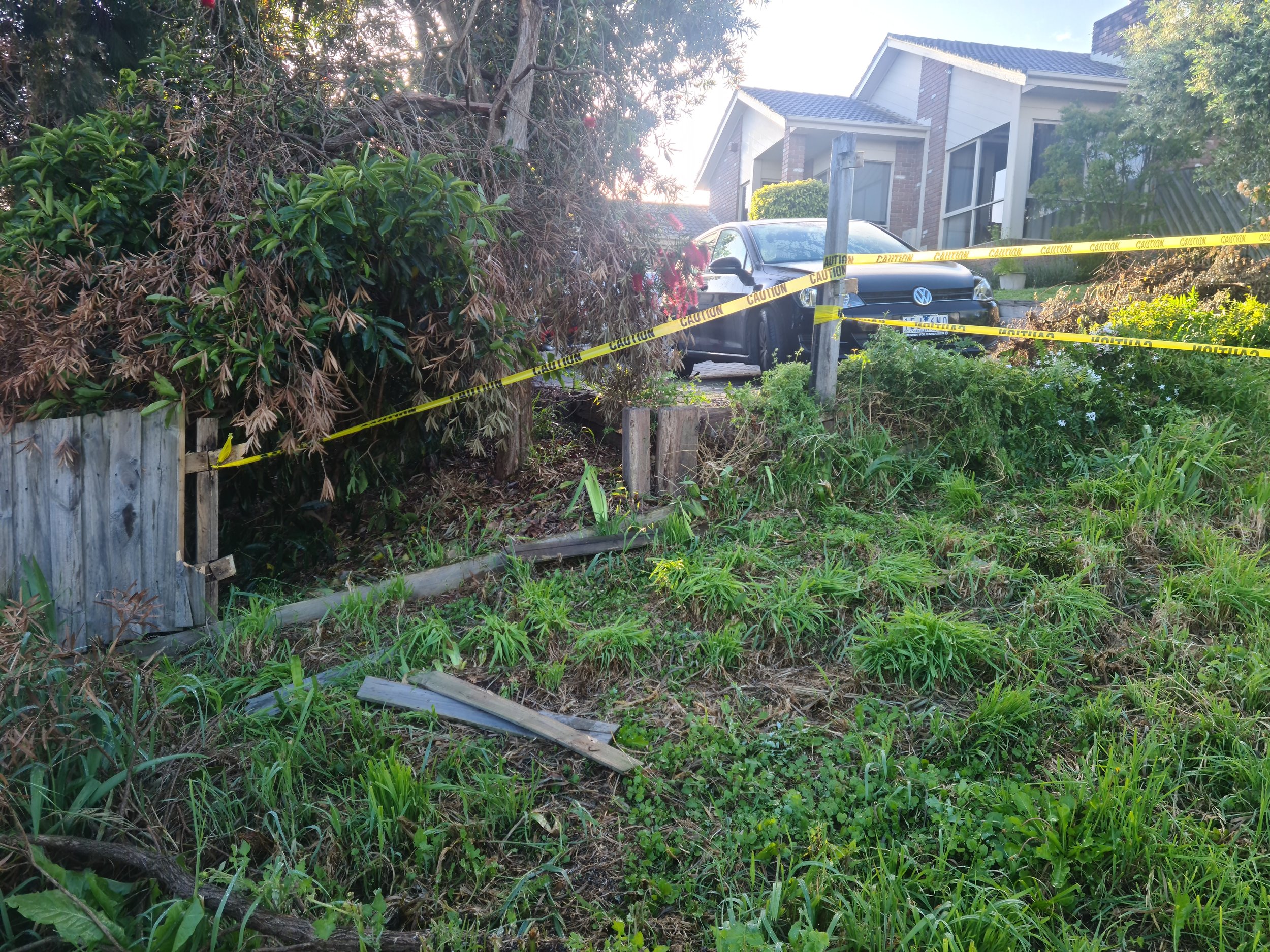 A residential yard with a damaged wooden fence, overgrown grass, and caution tape cordoning off the area, with a black Volkswagen car parked in front of a house in the background.
