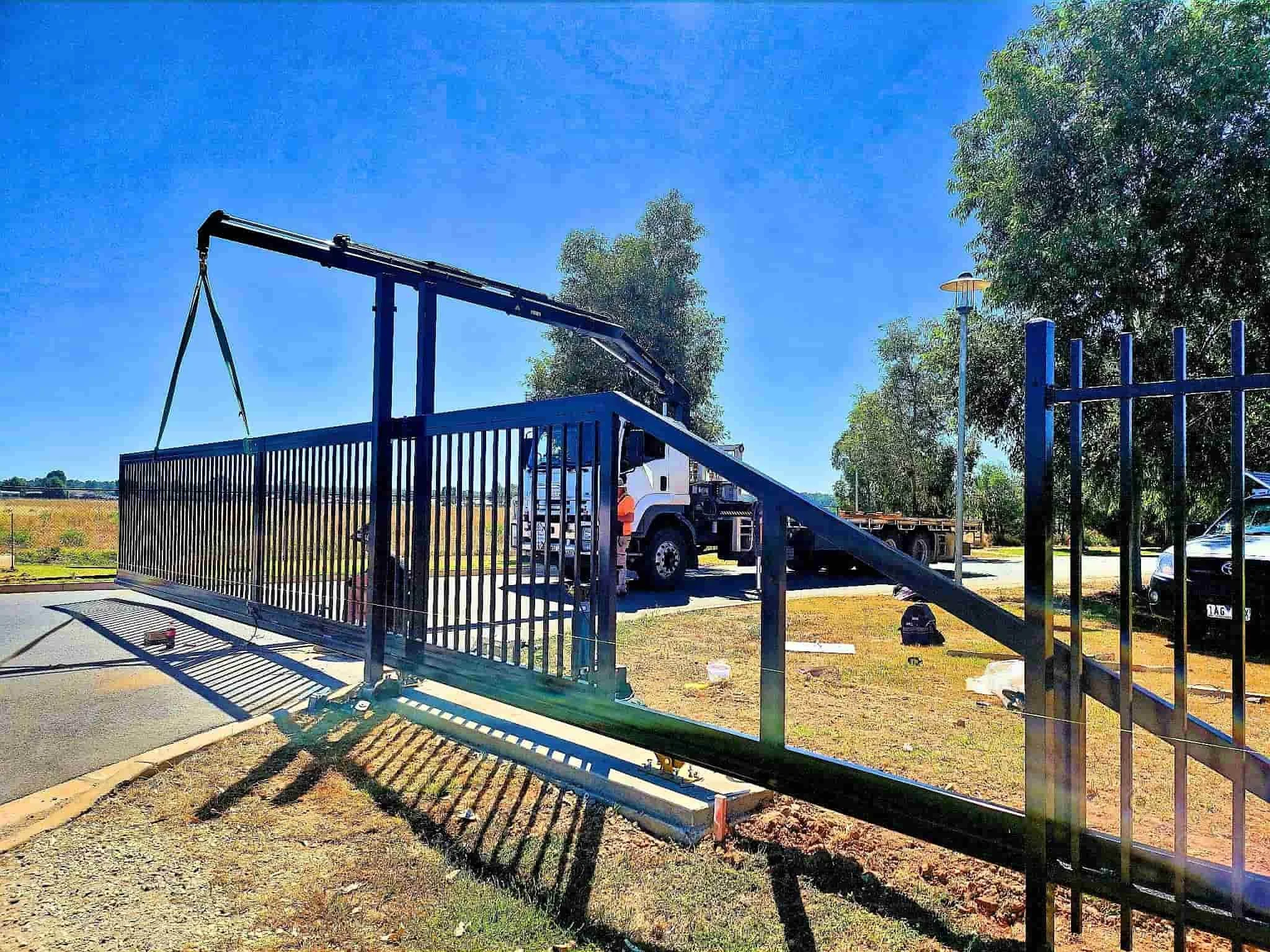 A large metal gate being installed with a crane, with trees and a parking lot visible in the background on a sunny day.