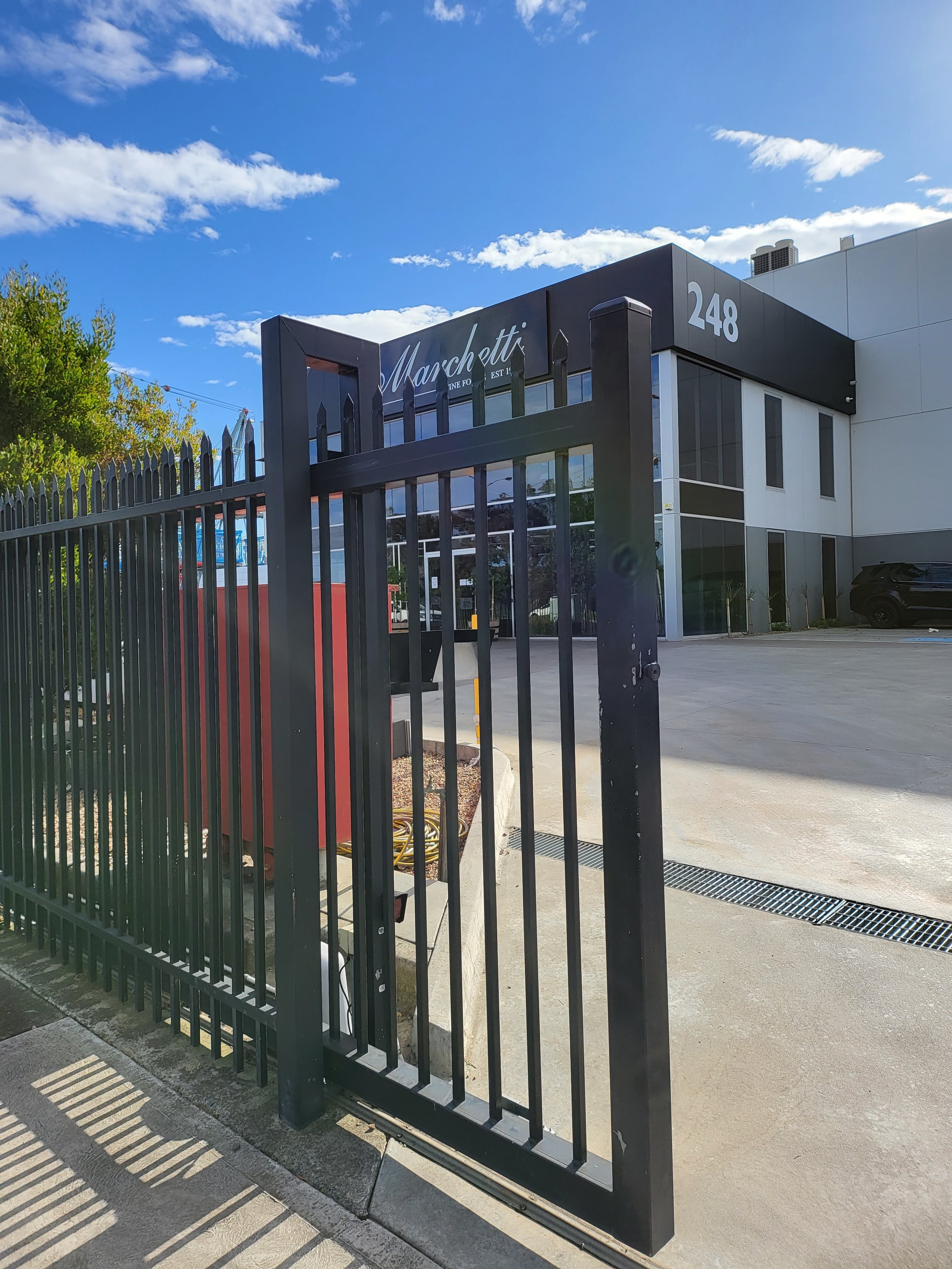 A black metal gate in front of a commercial building with the name 'Marschitti' and the number 248 on the facade. The building is modern with large windows and a parking lot. The sky is blue with some clouds.