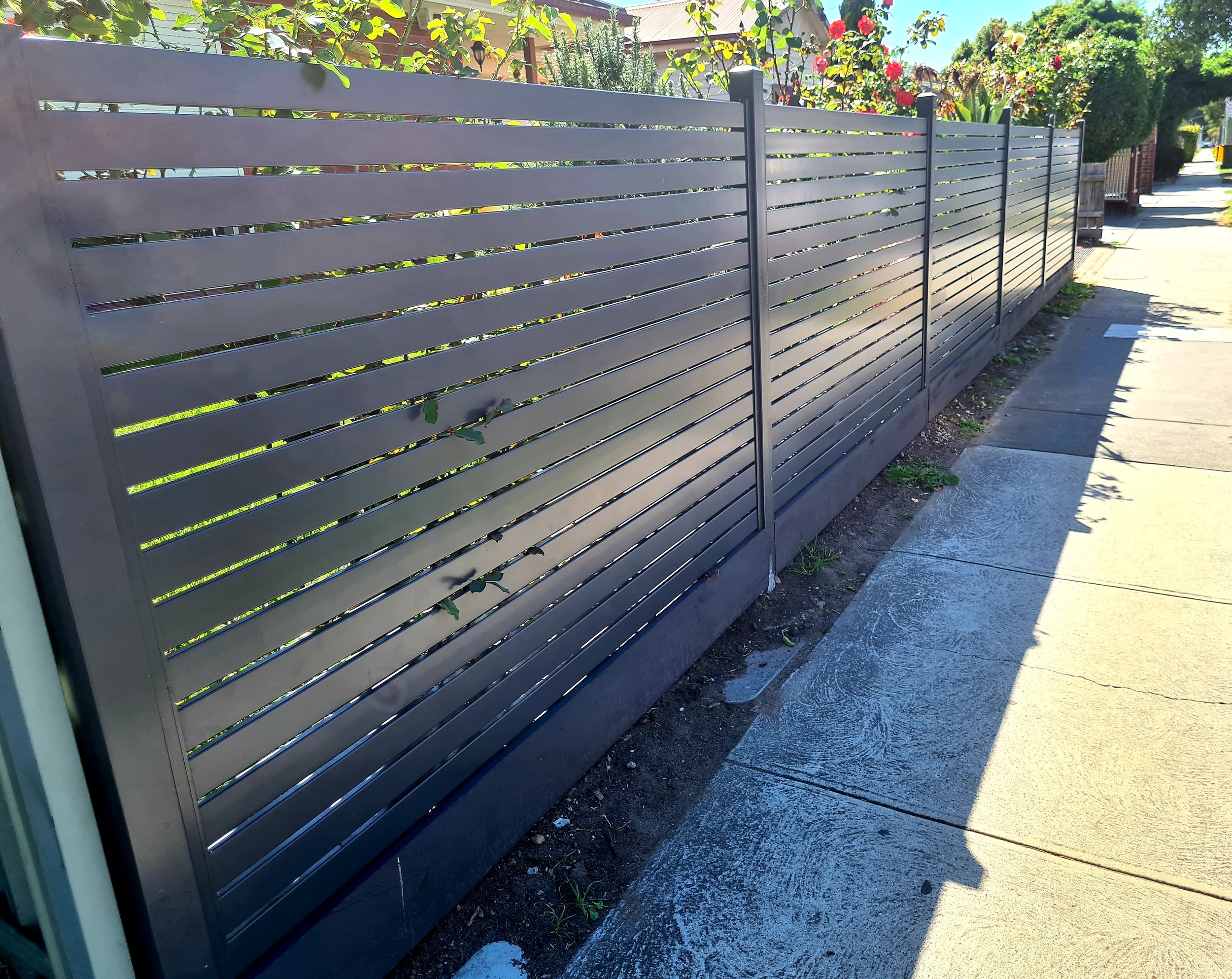 A modern horizontal slat metal fence along a sidewalk, with a garden of plants and flowers behind it and a sunny street scene.