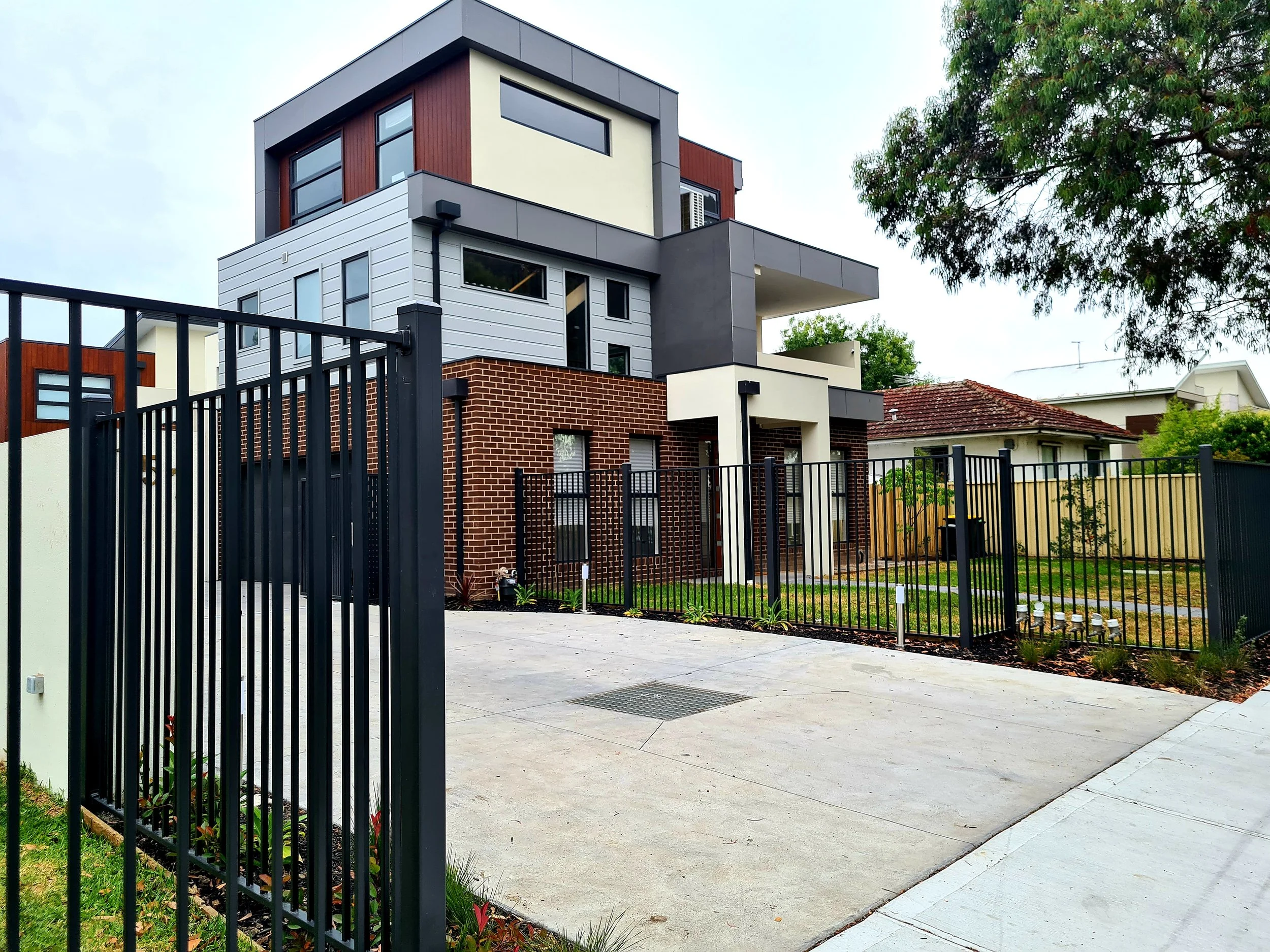 Modern multi-story house with brick and gray panel exterior, surrounded by a black metal fence, with a concrete driveway and a backyard with green grass and trees.