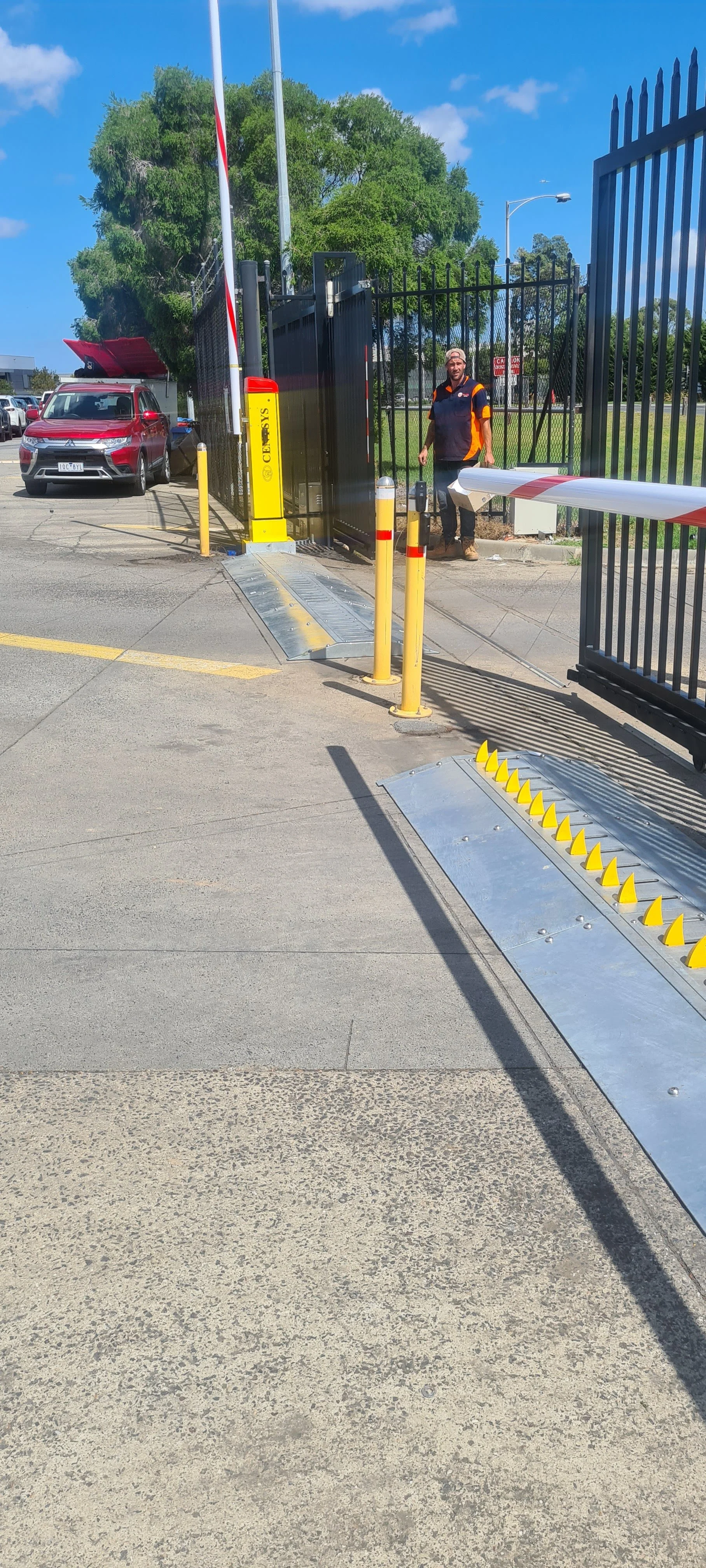 Entrance to a parking lot with a security gate, yellow safety barriers, a man in a dark uniform near the gate, and parked cars in the background.