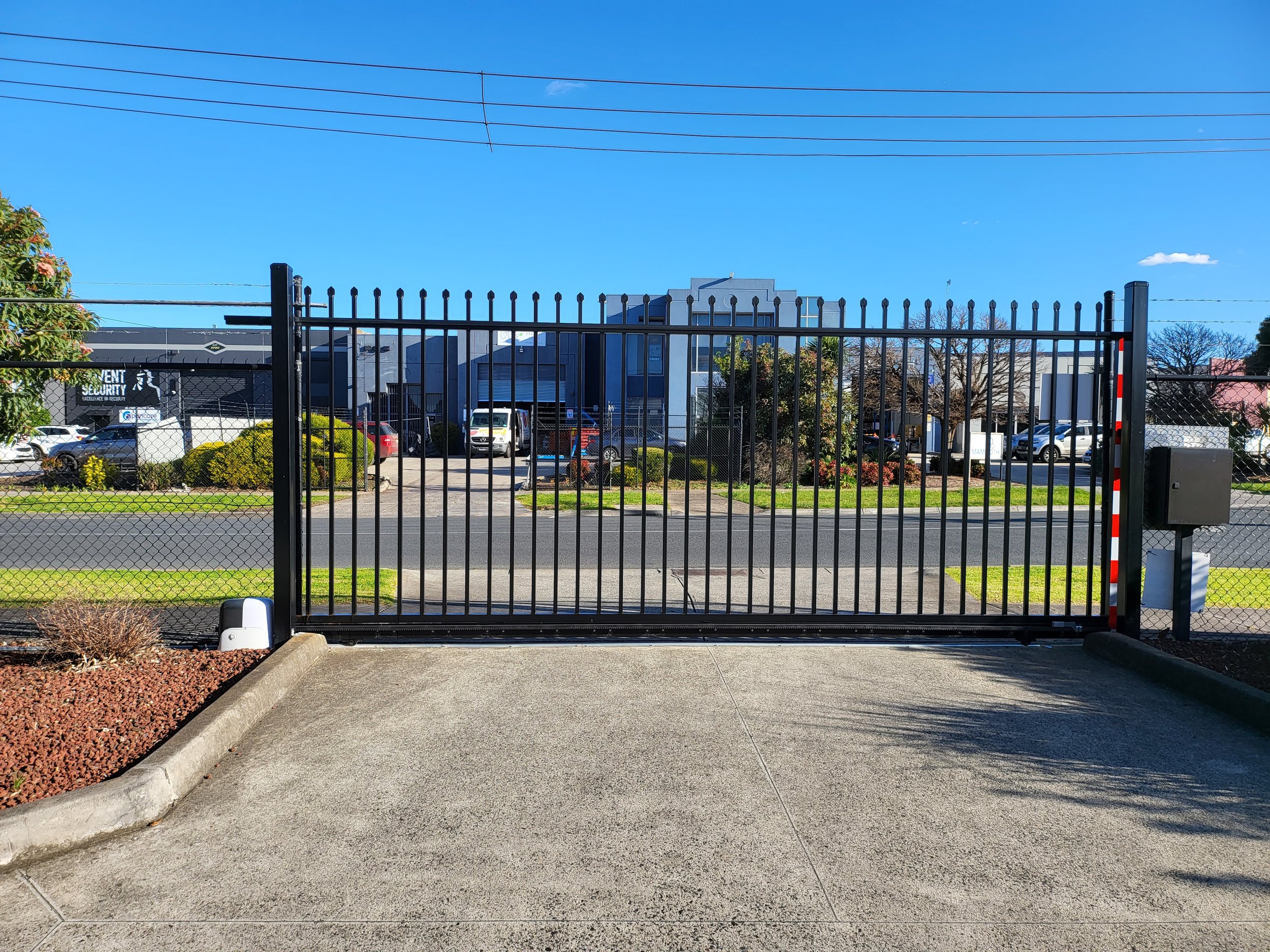 A black sliding security gate at the entrance of a commercial or industrial property, with a sidewalk and landscaped area in the foreground, and several vehicles and buildings behind the gate under a clear blue sky.