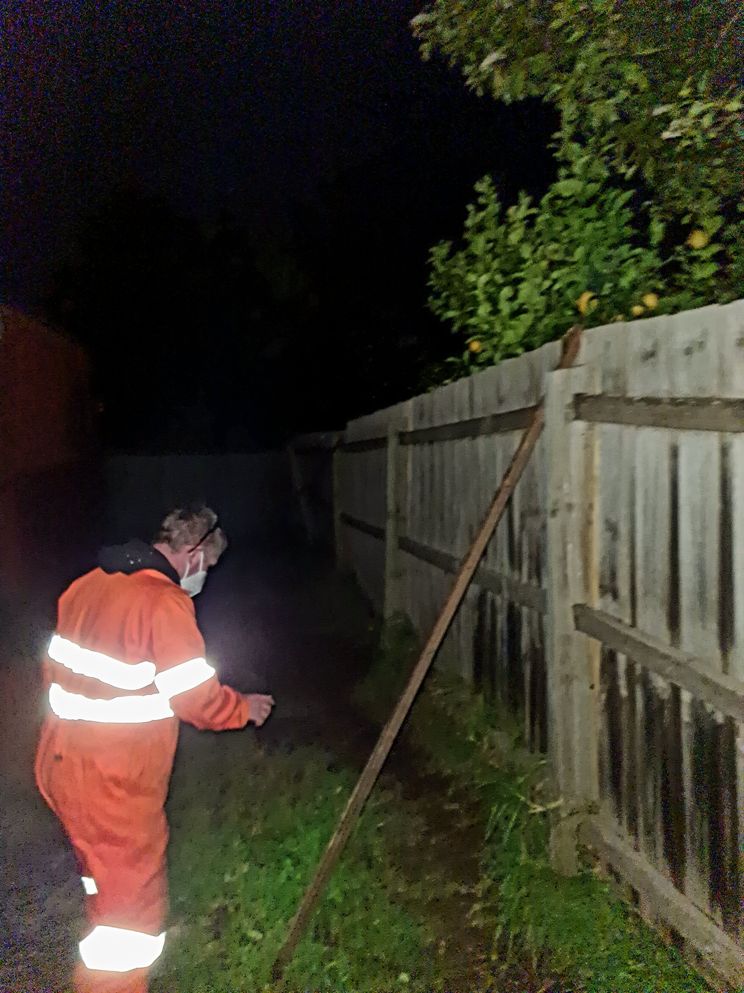 Person in an orange safety suit and face mask examining a fallen wooden fence post at night.