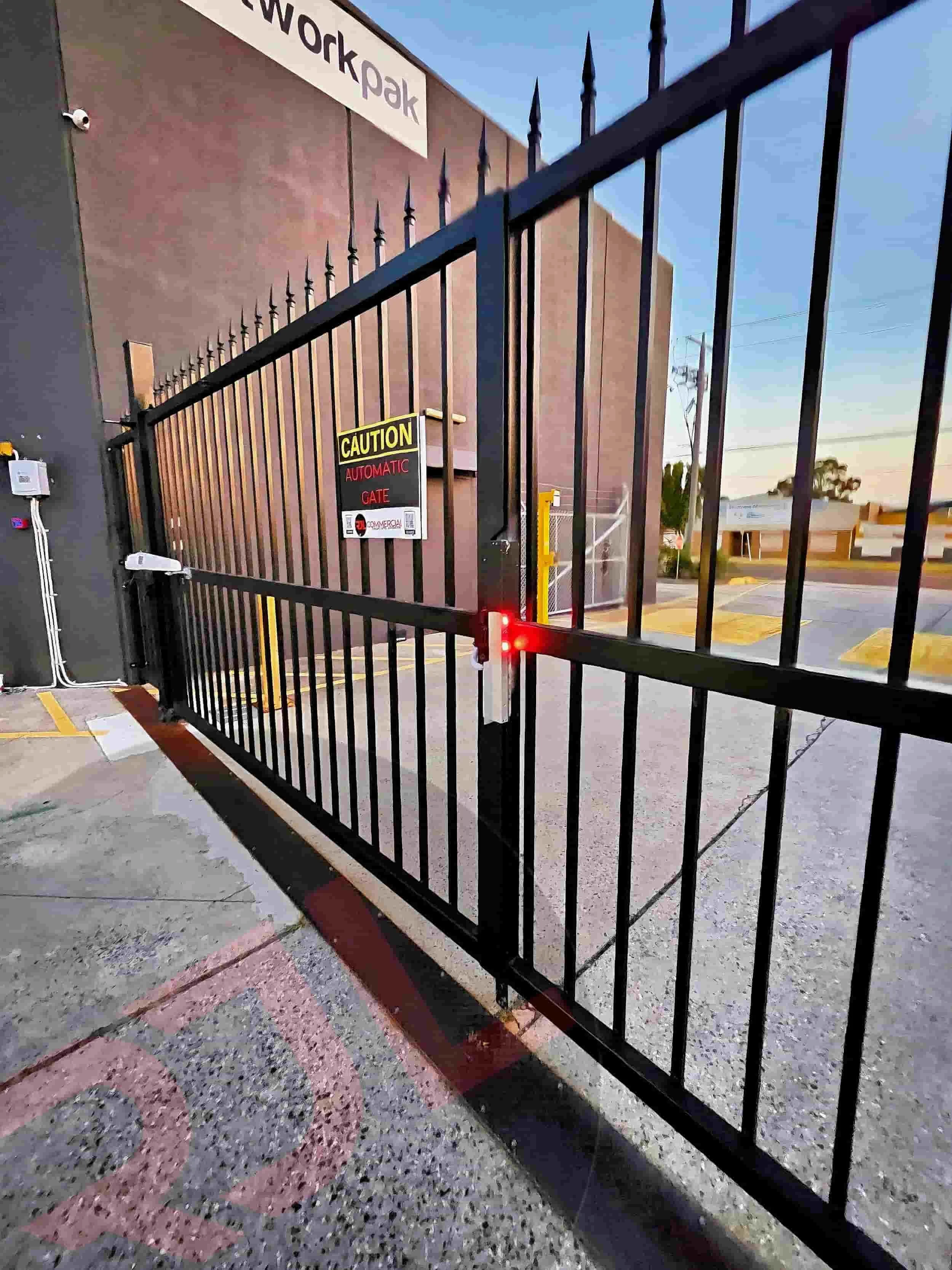 Black automatic gate with a yellow caution sign near a building with a sign that reads 'newpark'. The gate has red lights and is situated in a parking lot with marked parking spaces.
