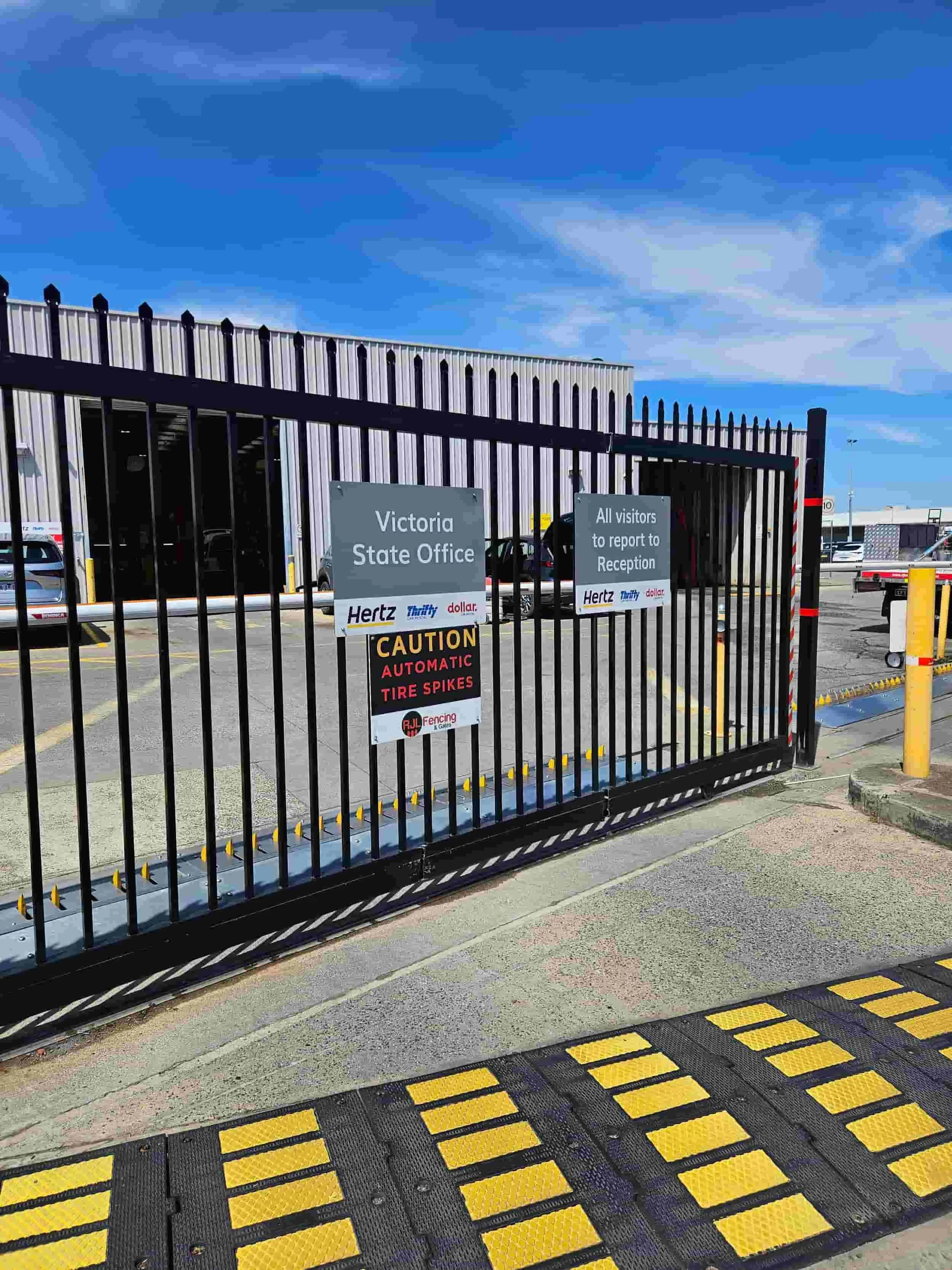 The entrance to the Victoria State Office parking lot, surrounded by a tall black metal fence with signs including caution for automatic tire spikes and directions for visitors; a parking lot with parked cars and a large industrial building in the ba