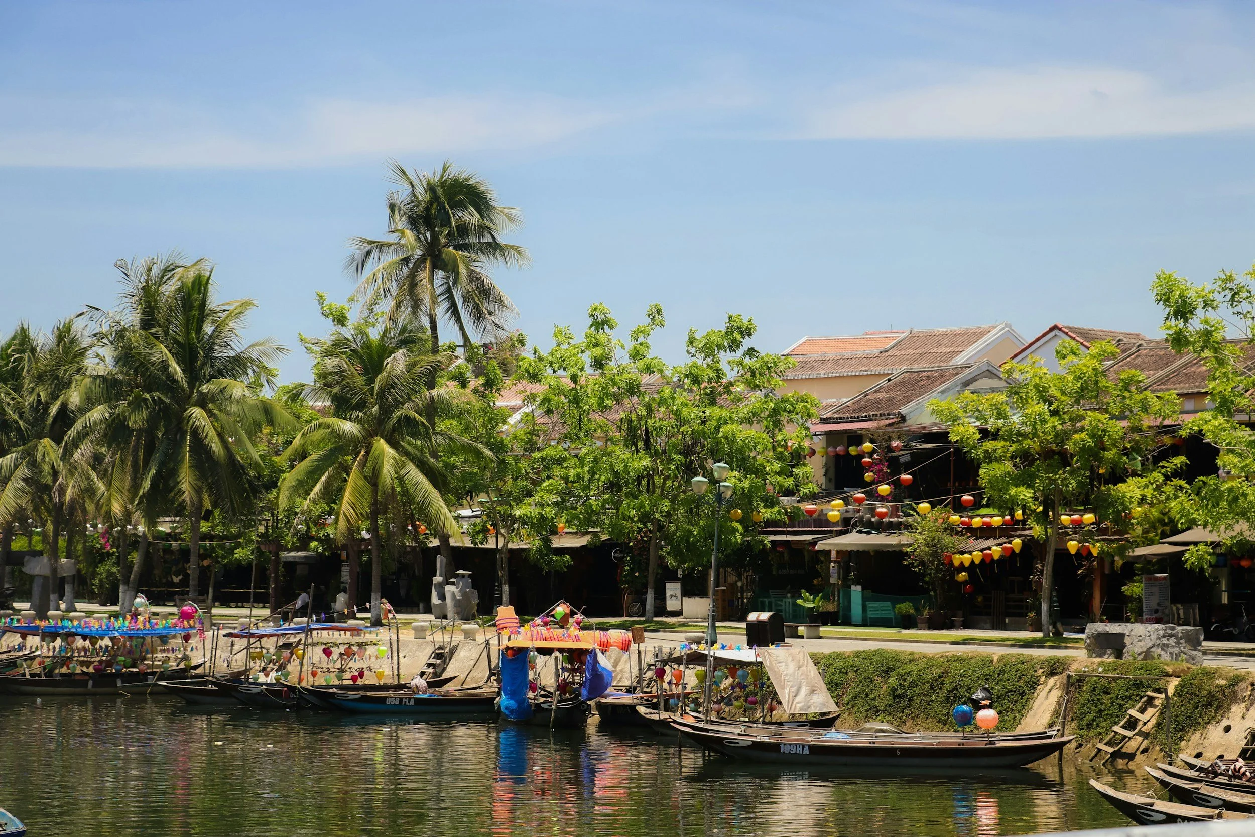 Boats floating on a river with trees and buildings decorated with colourful lanterns in the background on a sunny day - Hoian Town in Vietnam.