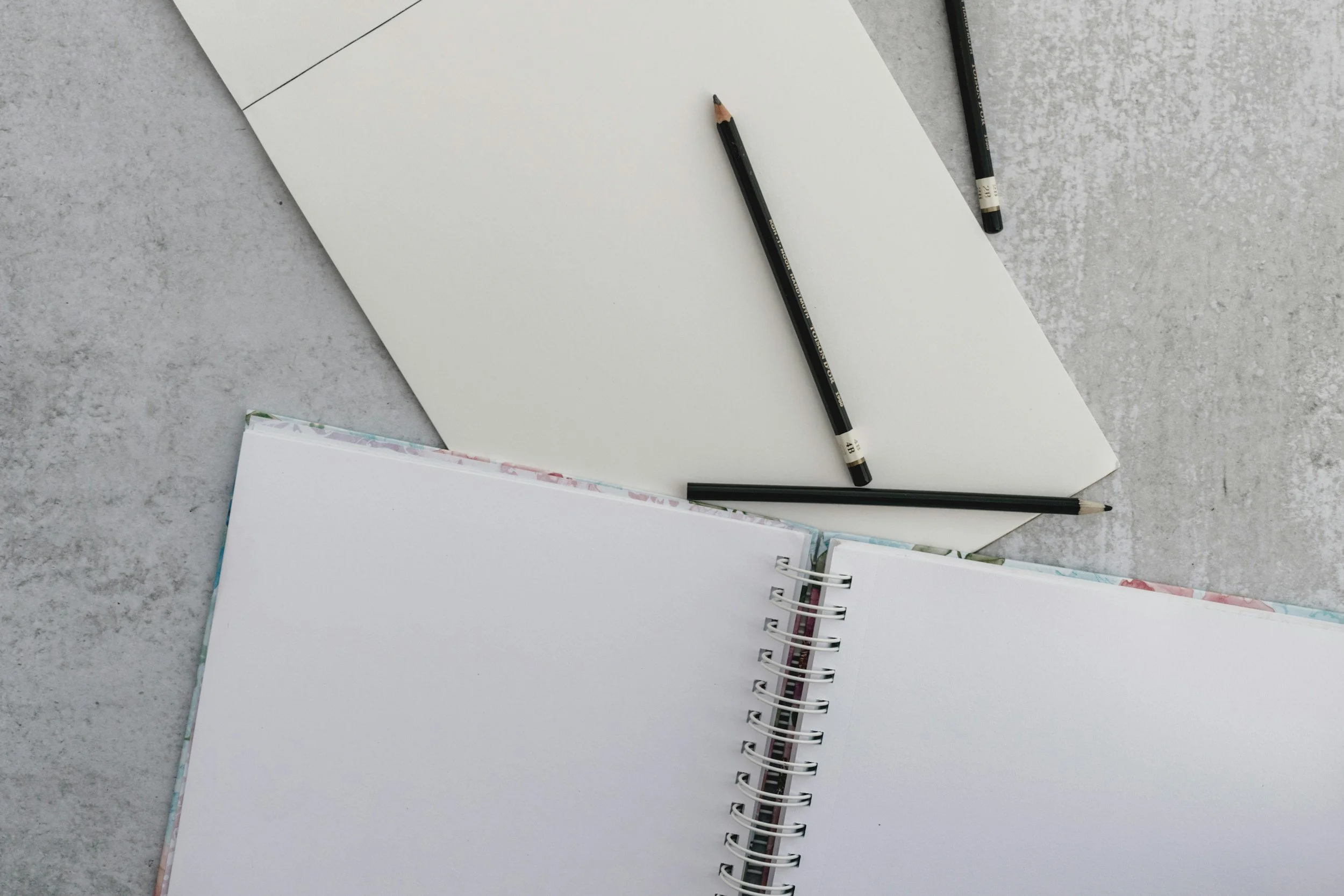 Open spiral notebook and blank sketchbook with black pencils on a gray surface.