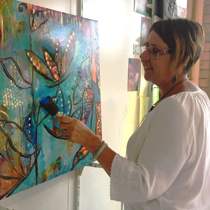 A woman with short hair and glasses smiling while holding a paint roller, painting a colourful, abstract artwork with floral designs on a canvas.