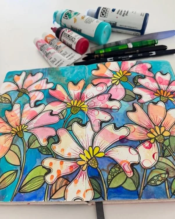 Colorful drawing of pink and white flowers with green leaves on a blue background, with art supplies including paint bottles, markers, and a pencil on a white surface.