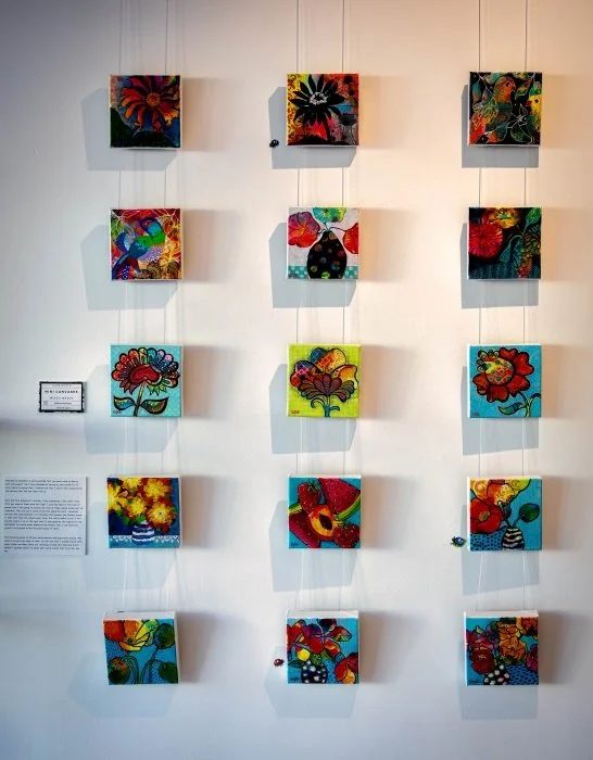 Nine colourful abstract paintings arranged in three rows of three on a white gallery wall, with small labels and an informational plaque on the left side.