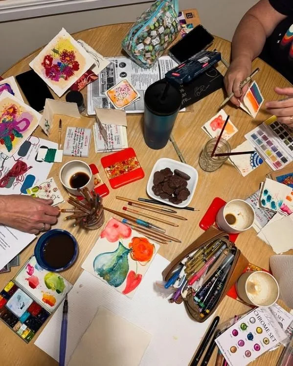 A group of people are sitting around a table engaged in painting and crafting activities, with various art supplies, brushes, paints, cups of coffee, and finished and in-progress artwork visible on the table.