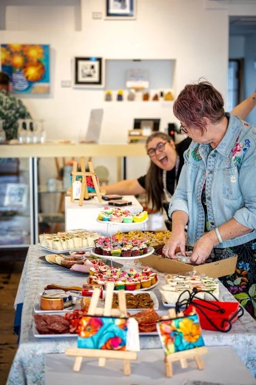 Two women at a table with a variety of colorful baked goods, cupcakes, and treats, in a room decorated with artwork and shelves.