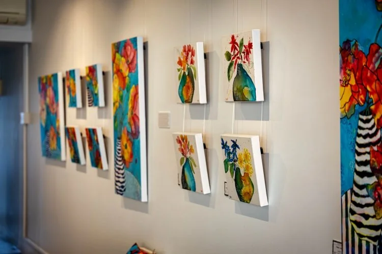 Colorful floral paintings displayed on a gallery wall, some mounted on white panels.