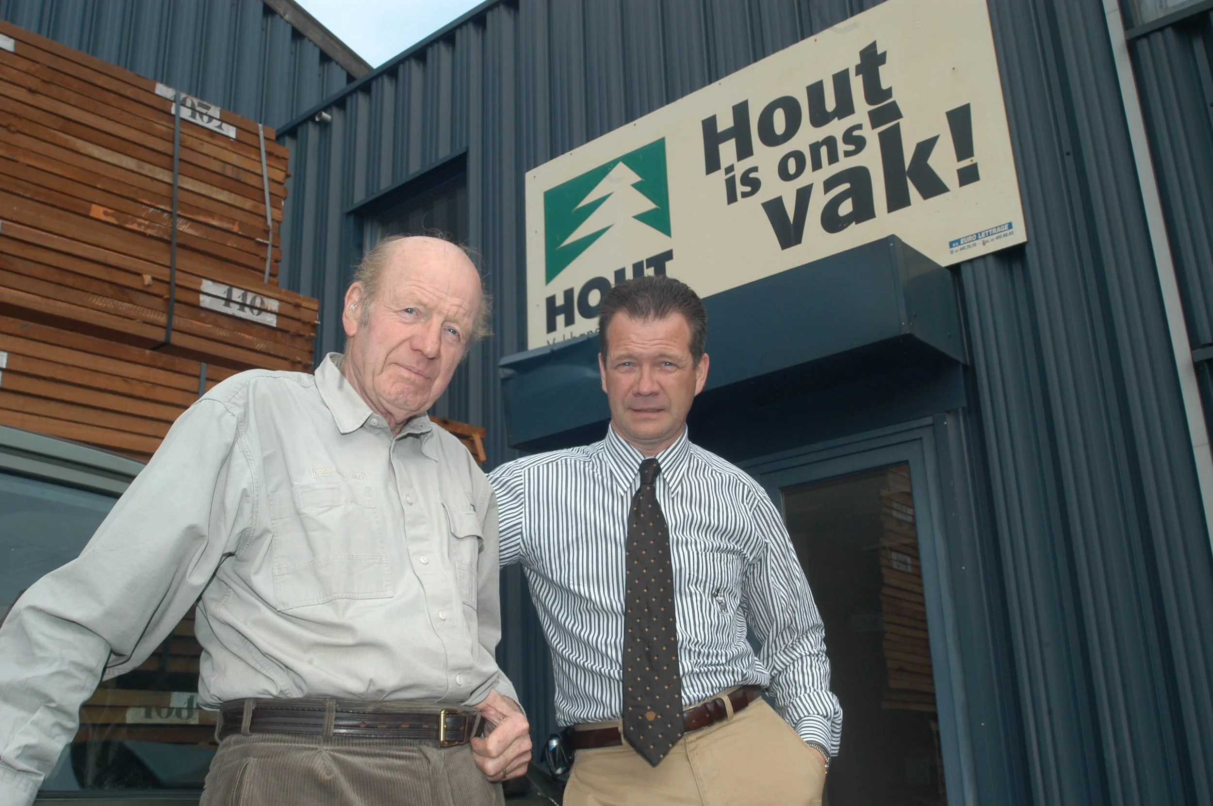 Twee mannen staan voor een houtwinkel die een groot bord met de tekst 'Hout is ons vak!' heeft, met stapels hout achter hen.