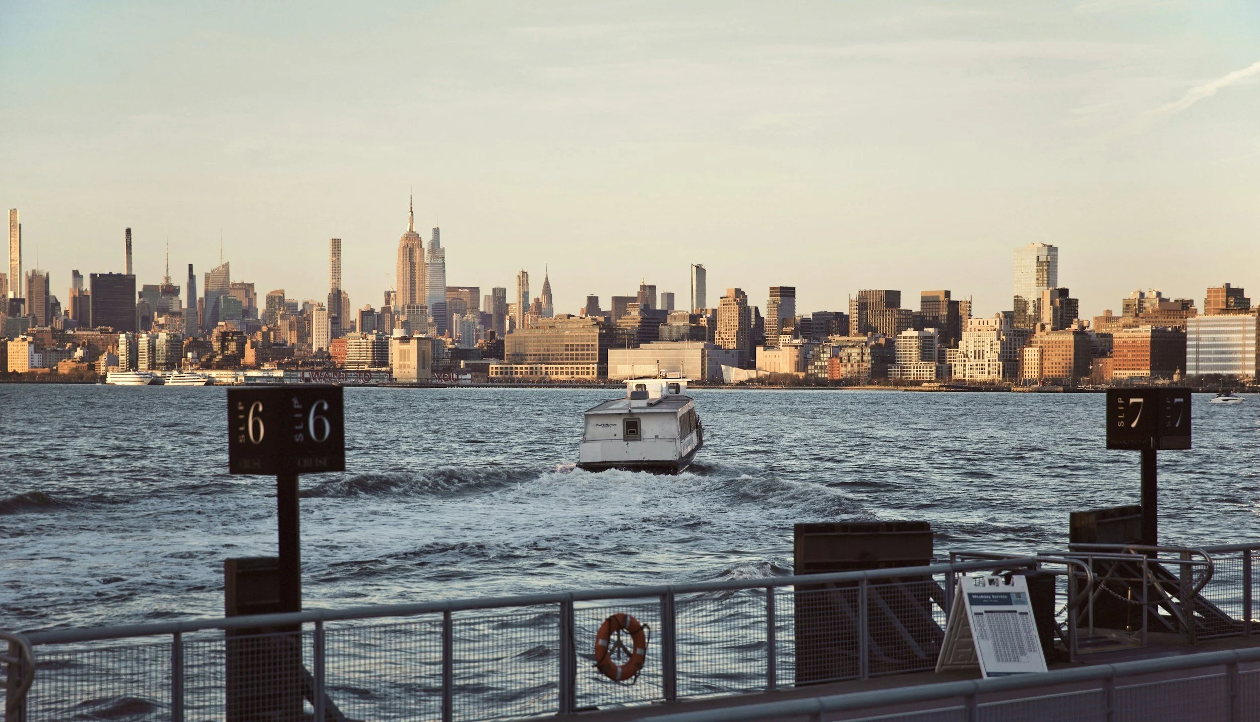 View of Manhattan skyline from waterfront with a boat in the water, and sailing docks in the foreground.