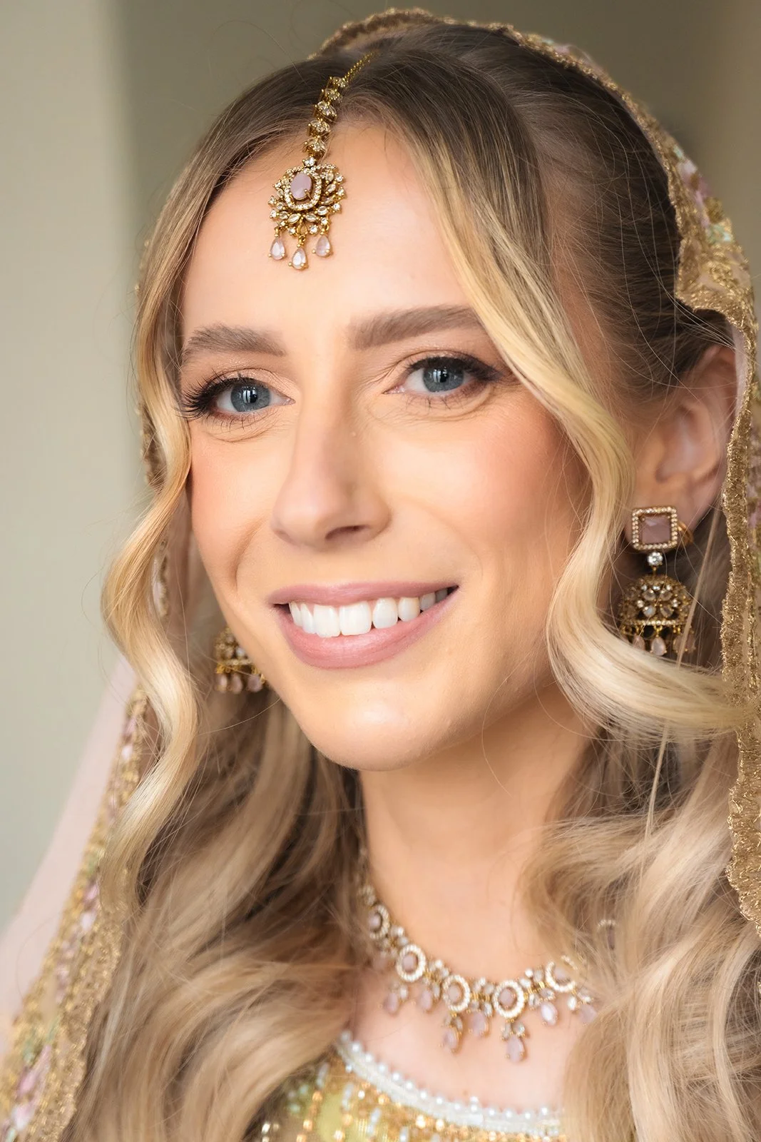 A bride dressed in traditional South Asian wedding attire, wearing elaborate jewelry including earrings, a necklace, and a headpiece, with styled blonde hair and light makeup, smiling.