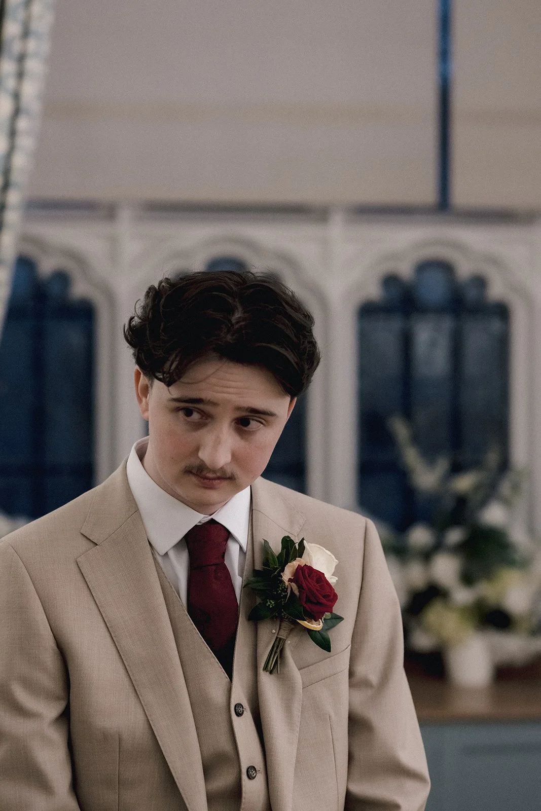 A groom with dark, curly hair and a mustache, dressed in a beige wedding suit with a white shirt and red tie, stands indoors with a pensive expression. He has a boutonniere with red and white roses pinned to his lapel.