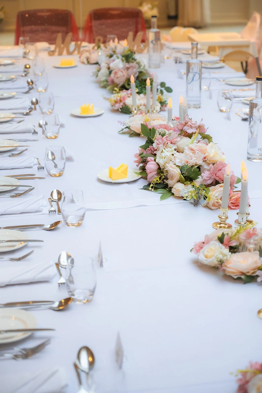 A long wedding banquet table decorated with pink and white floral arrangements, white candles, and place settings with plates, silverware, and glasses, set for a formal event or wedding reception.