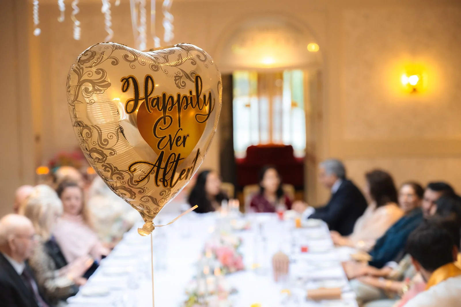 A heart-shaped balloon with the words "Happily Ever After" floats in the foreground, with a group of people sitting at a long table in a decorated indoor venue in the background.