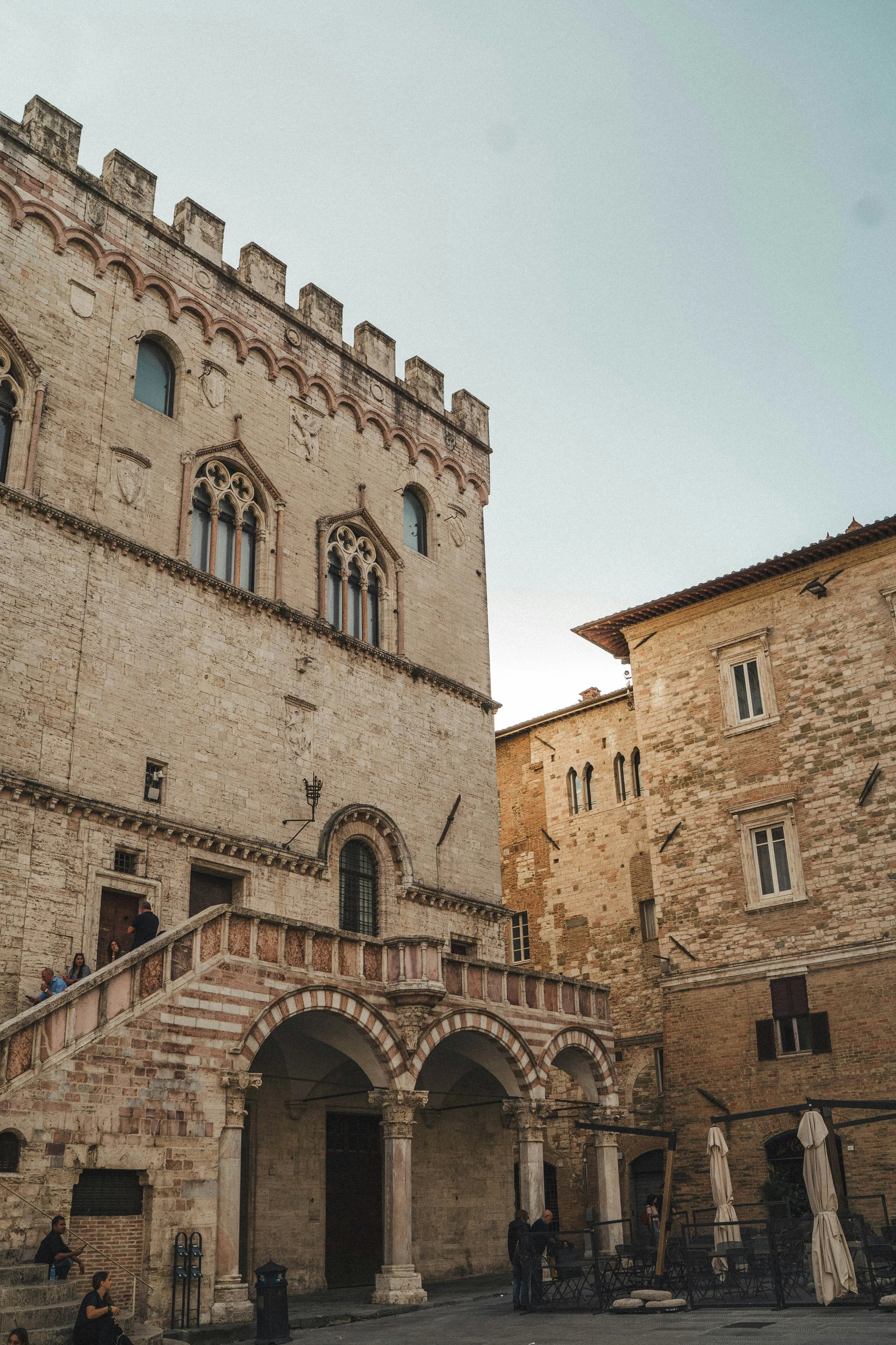 Historic stone building with gothic arched windows and an elevated walkway supported by columns, with people sitting and walking in the courtyard.