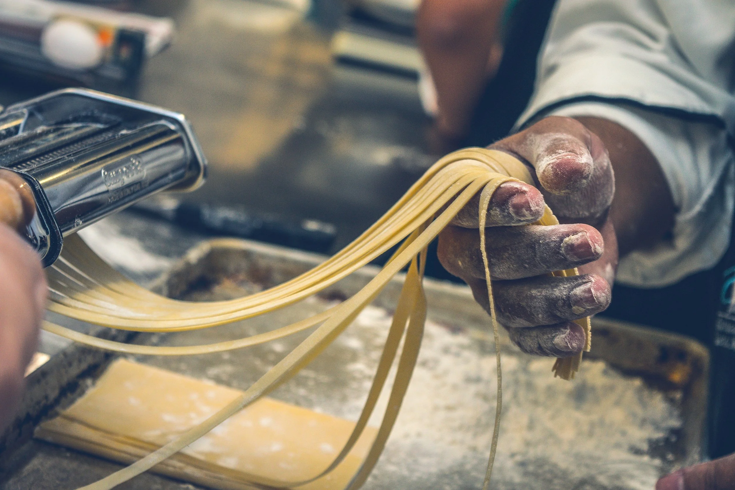 Close-up of a person's hand holding uncooked pasta noodles, with a pasta machine in the background, dusted with flour.