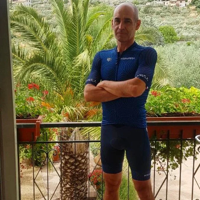 A man in cycling gear standing with arms crossed on a balcony with lush green plants and trees in the background.