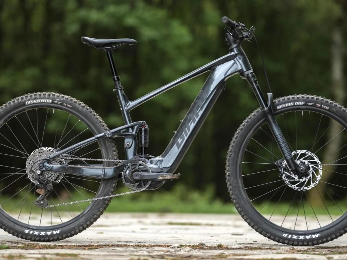A black full-suspension mountain bike resting on a wooden surface with a blurred green forest background.