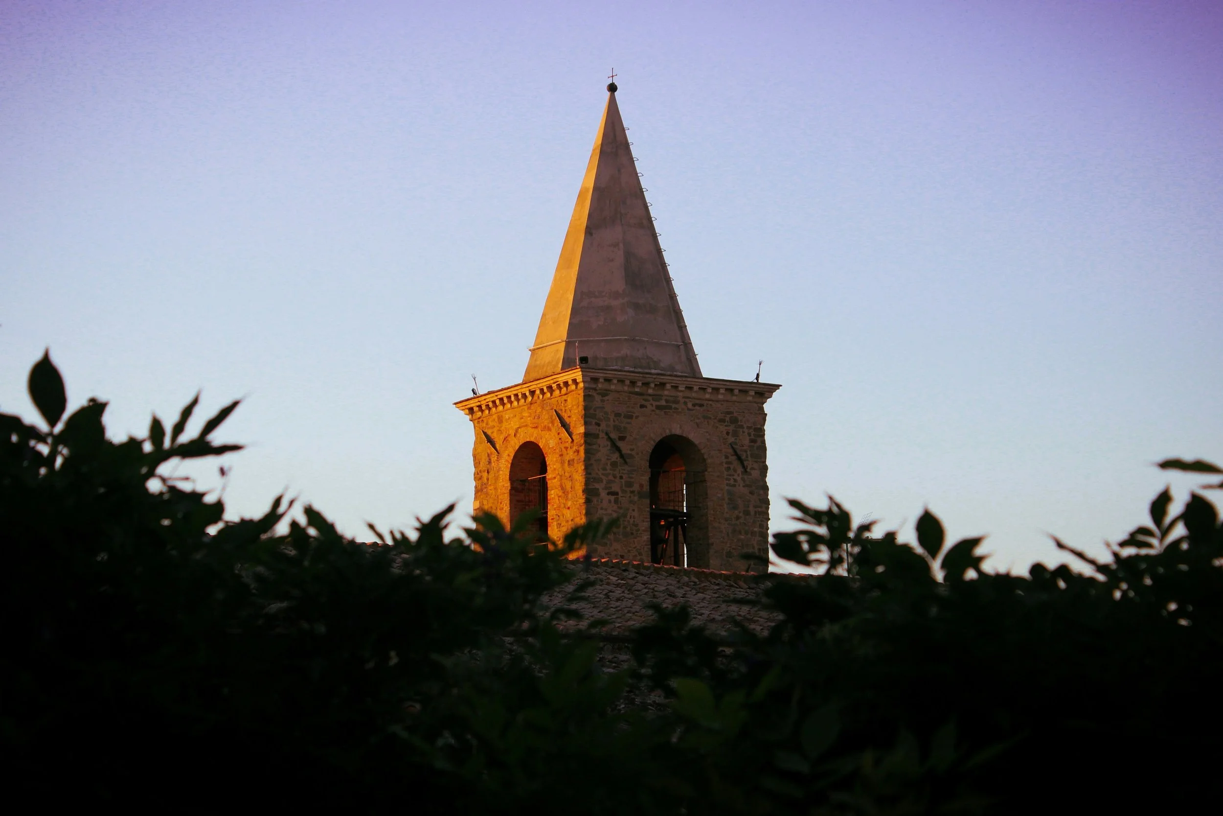 A stone church tower with a sharp spire, illuminated by golden light, seen through dark foliage against a pale blue sky.