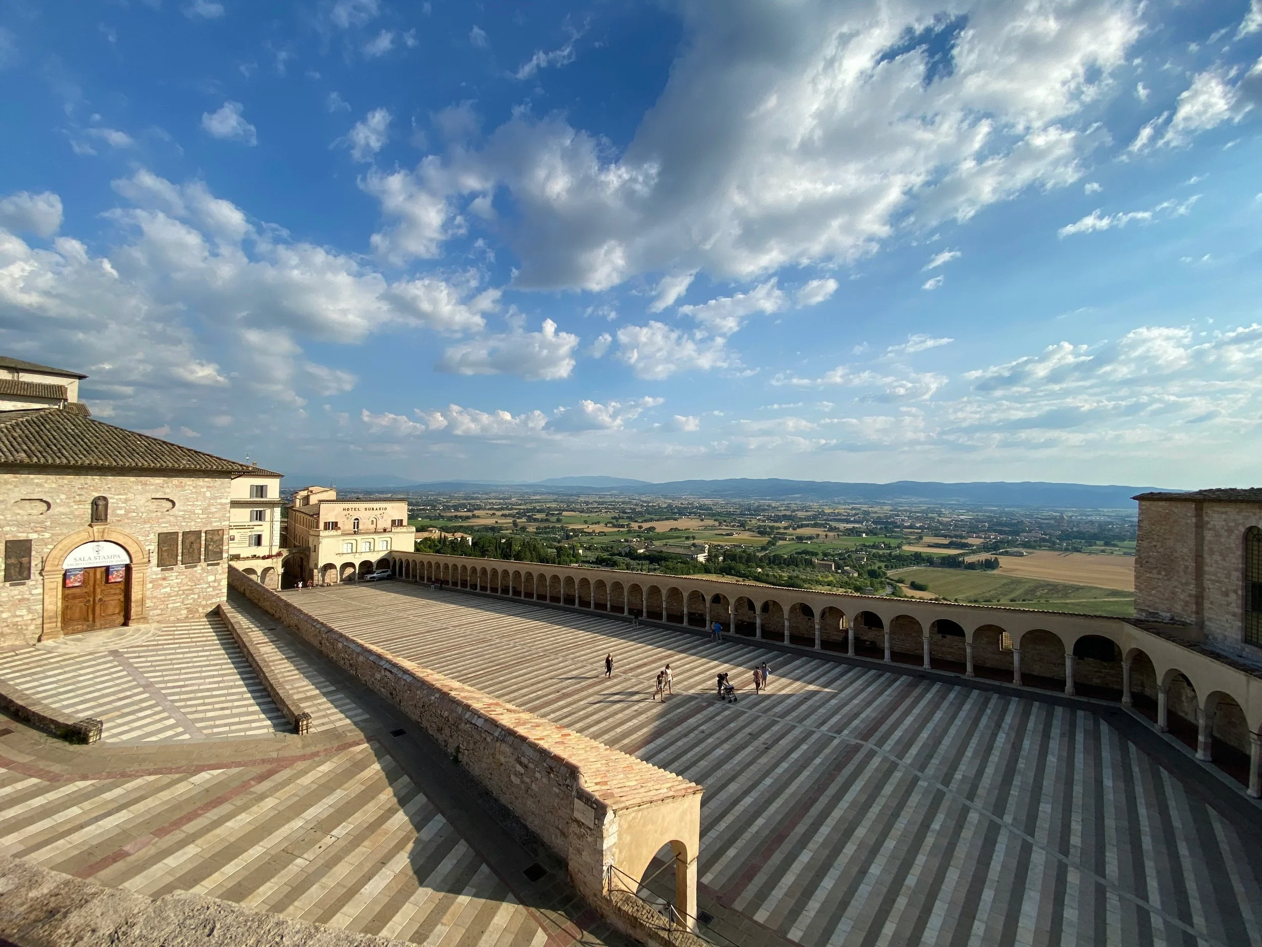 A panoramic view of a historic stone plaza with striped pavement and an arched colonnade under a blue sky with scattered clouds, overlooking a green and farmland landscape.