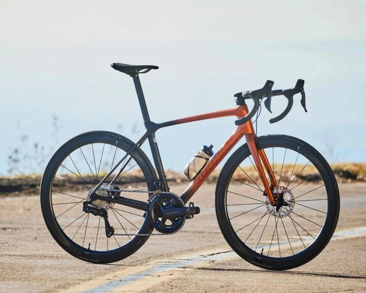 A sleek modern road bicycle with a two-tone black and orange frame, thin tires, water bottle, and drop handlebars, parked outdoors on a paved road near a body of water.