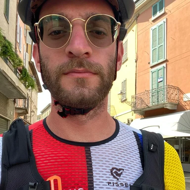 Close-up of a man wearing vintage sunglasses and a cycling jersey, standing outdoors in a European city with colorful buildings and balconies.