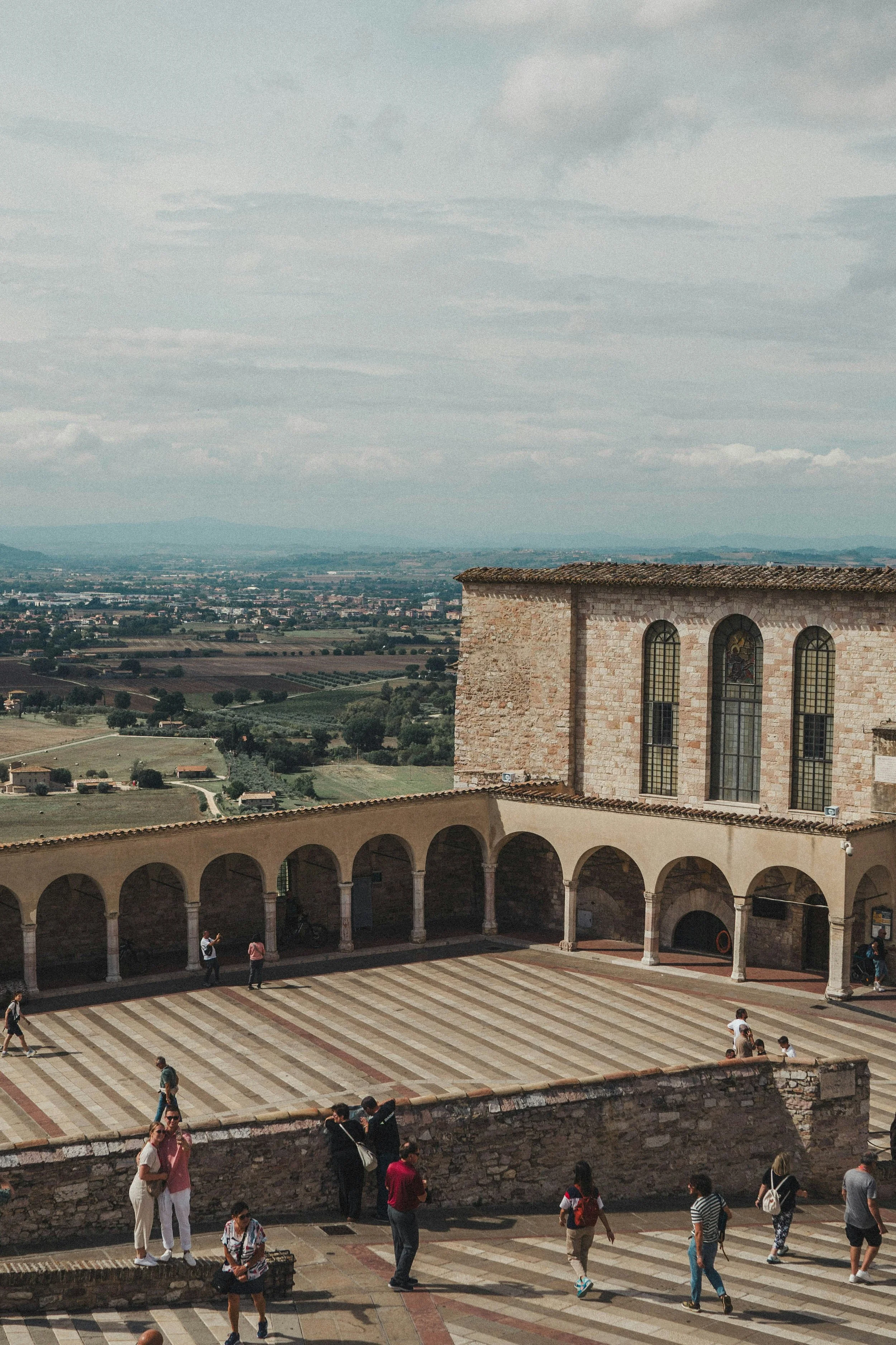 People walking in Assisi, on a stone courtyard outside an old stone church San Francesco with large arched windows, surrounded by a colonnade and overlooking a rural landscape, perfect for cycling with rental bike
