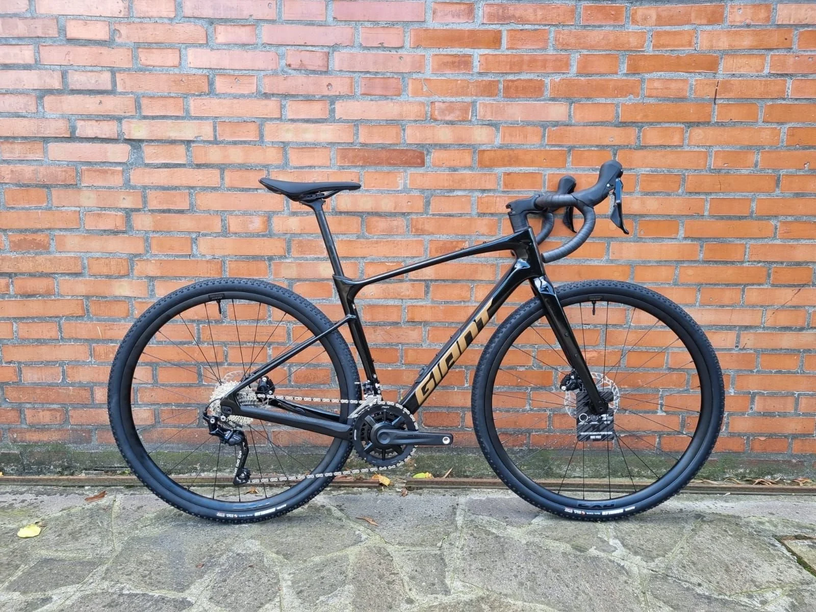 A black and tan road bicycle leaning against a brick wall on a paved surface.