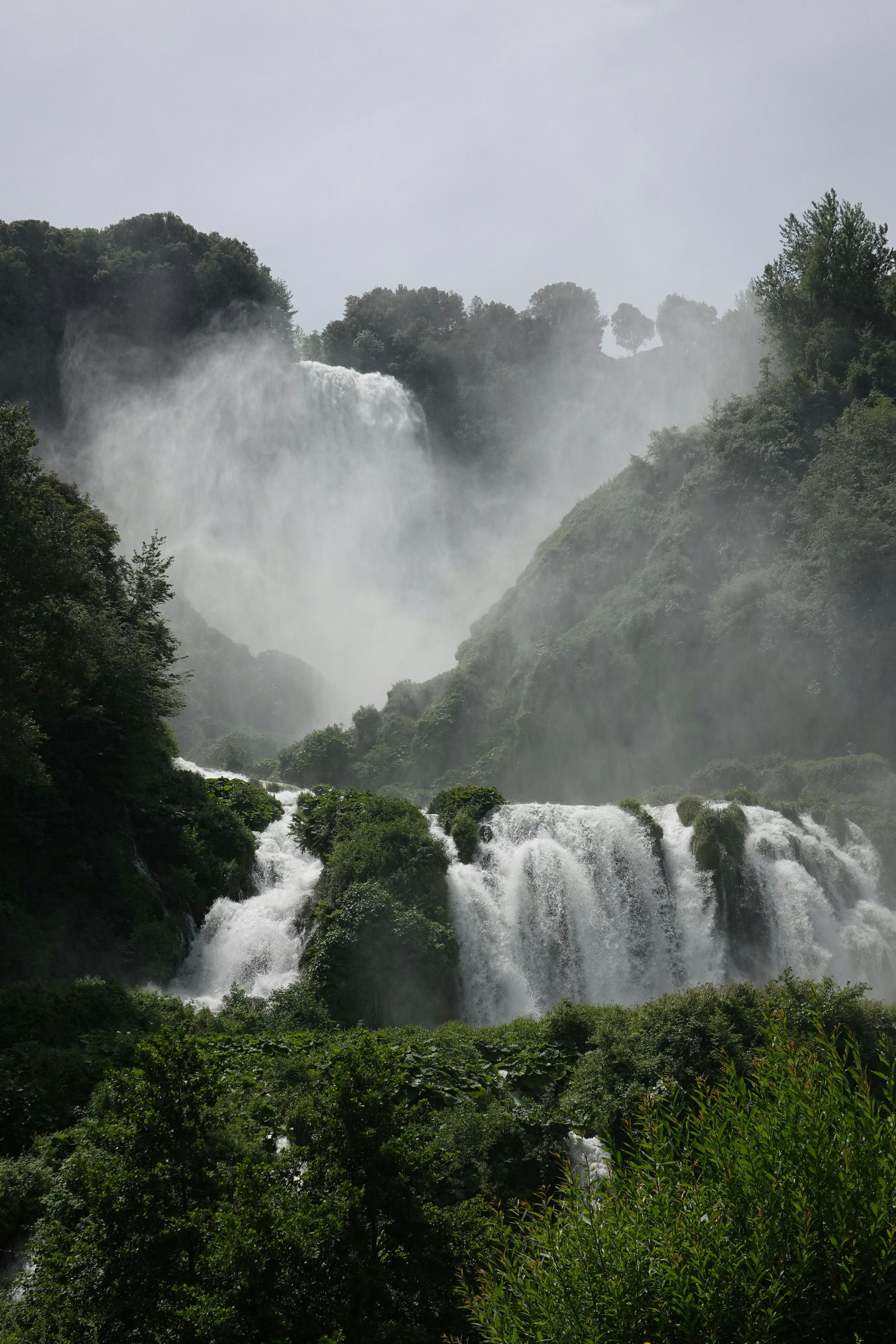 Tall waterfall cascading down green rocky cliffs surrounded by lush trees and mist in a natural landscape.