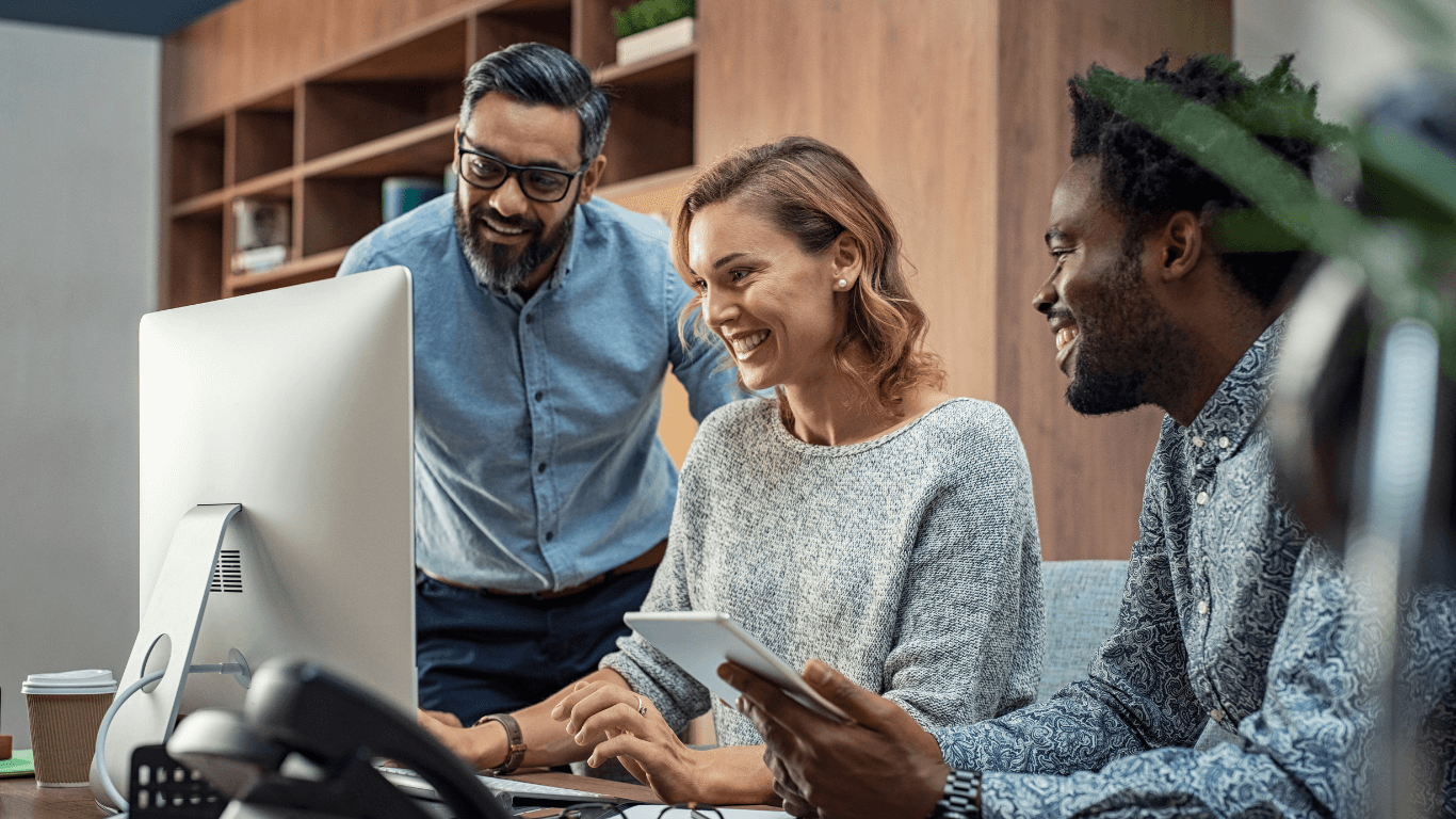 Three colleagues gathered around a computer, looking at the screen and smiling in an office setting.