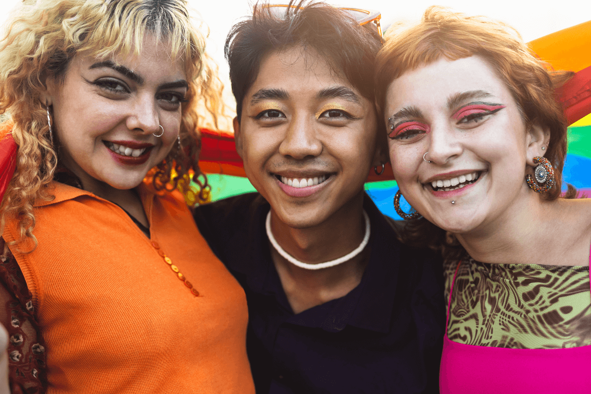 Three joyful individuals smiling together at an outdoor pride event, with rainbow flags in the background