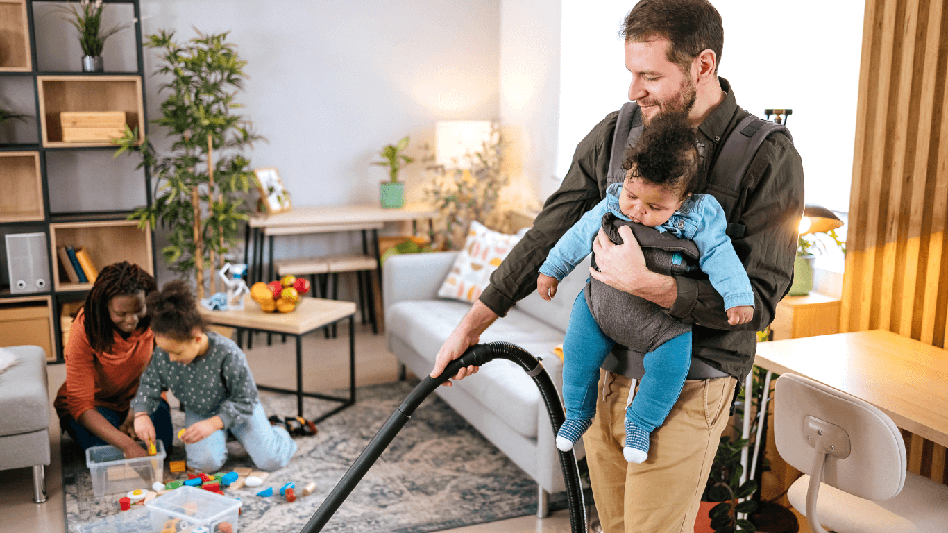A man vacuuming with a young child in a baby carrier on his chest, while two children play with toys on the floor in a cozy living room.